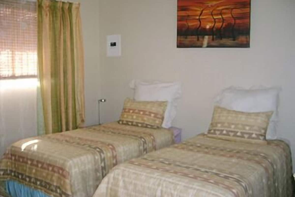 A room with two beds, each with patterned bedding and pillows. There is a window with curtains, a painting on the wall, and a small table with a lamp.