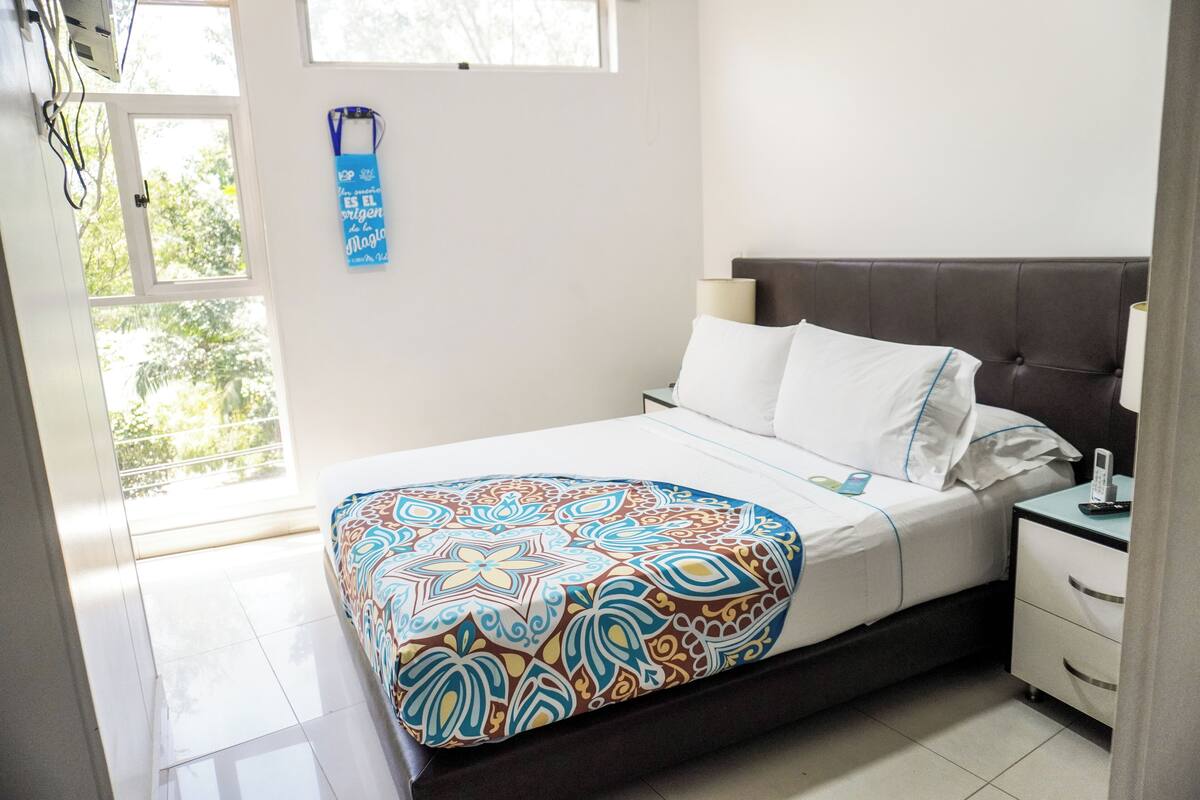 A hotel room with a bed, a nightstand, a window, and a door.