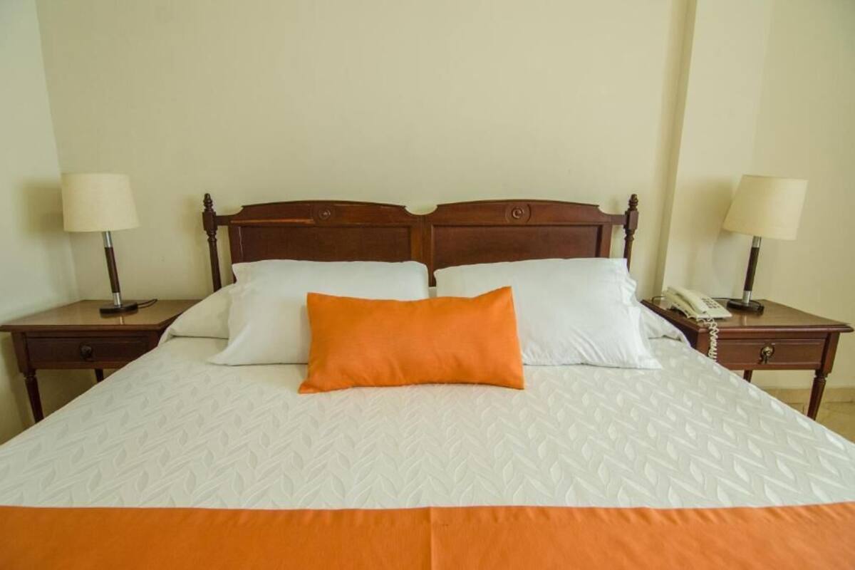 Una cama bien tendida con una colcha estampada, dos almohadas y una almohada anaranjada decorativa. Se ven dos mesitas de noche, una de ellas con una lámpara y un teléfono.