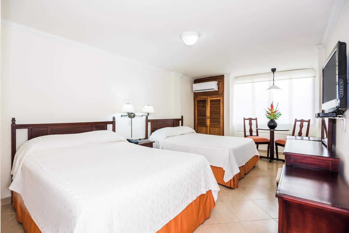 Habitación de hotel con dos camas, un escritorio con televisor, una mesita con sillas y una ventana con cortinas.