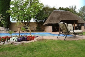 A pool area with a thatched-roof structure, a lounge chair, and colorful flower beds. - Augusta Villa St Pio Guest House (Edenvale)