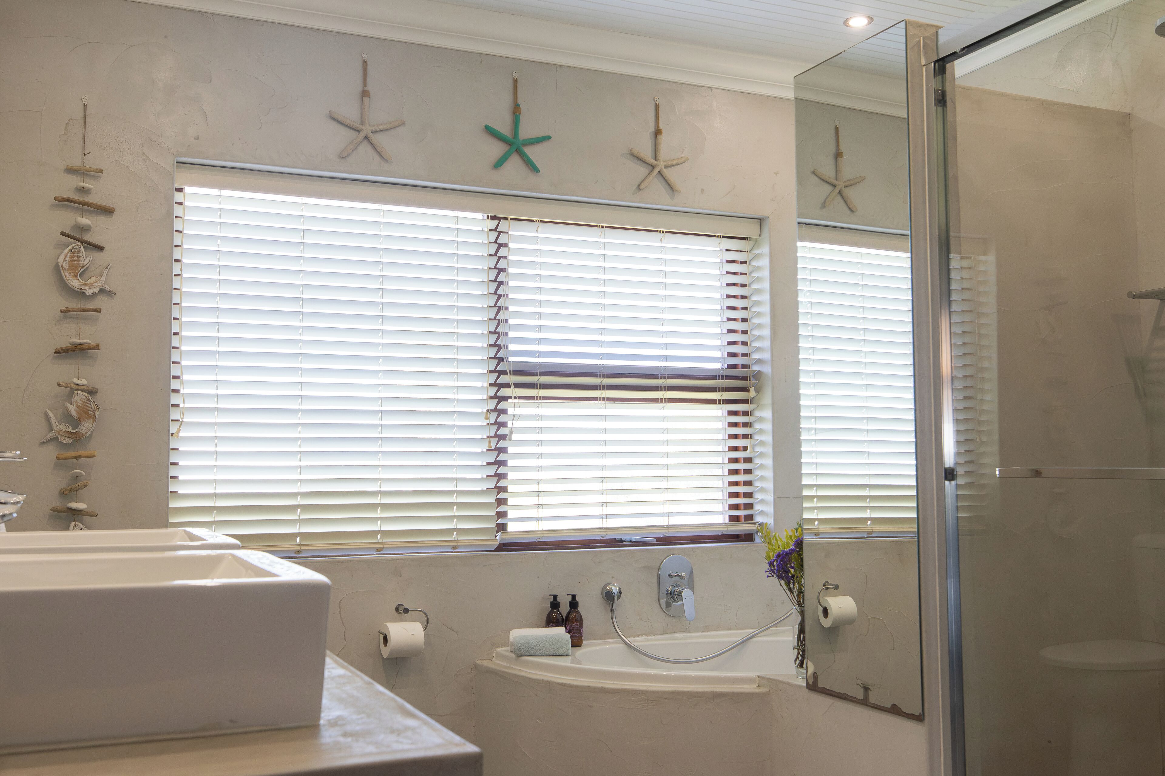 Ein Badezimmer mit Badewanne, Waschbecken und Dusche. An der Wand sind Dekorationen mit Seesternen angebracht und ein Fenster mit Jalousien.