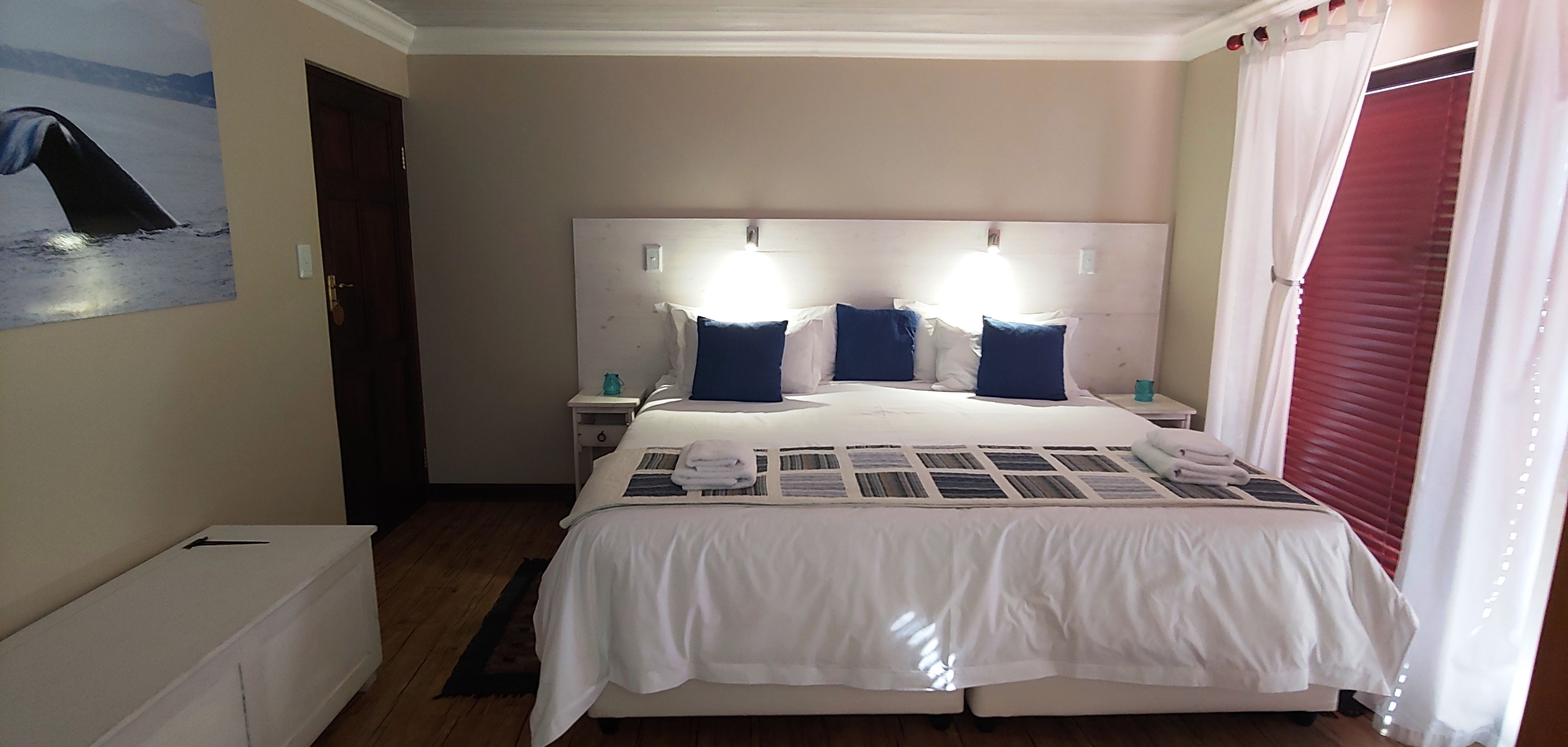 A neatly arranged hotel room with a large bed, two bedside tables, a nightstand, a wall-mounted lamp, and a window with red blinds.