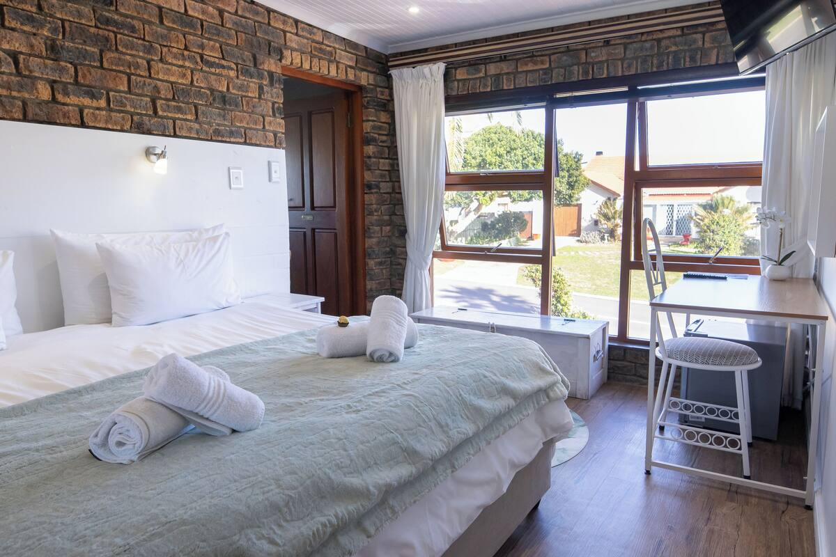 A hotel room with a brick wall, a large window with curtains, a bed with white linens, and a small desk with a chair.