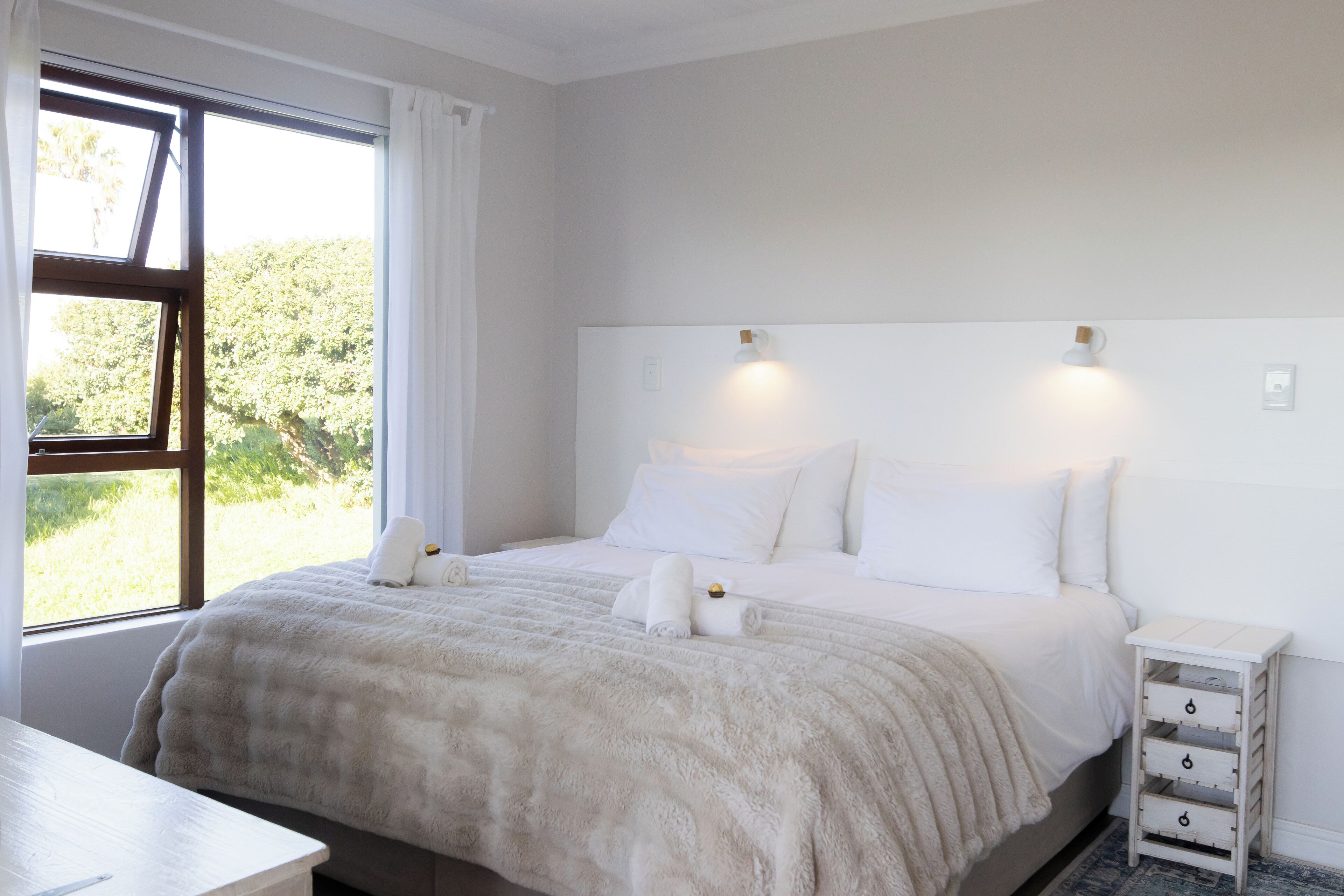 A neatly made bed with white bedding and a fluffy white throw blanket, a bedside table with drawers, and a window with curtains.