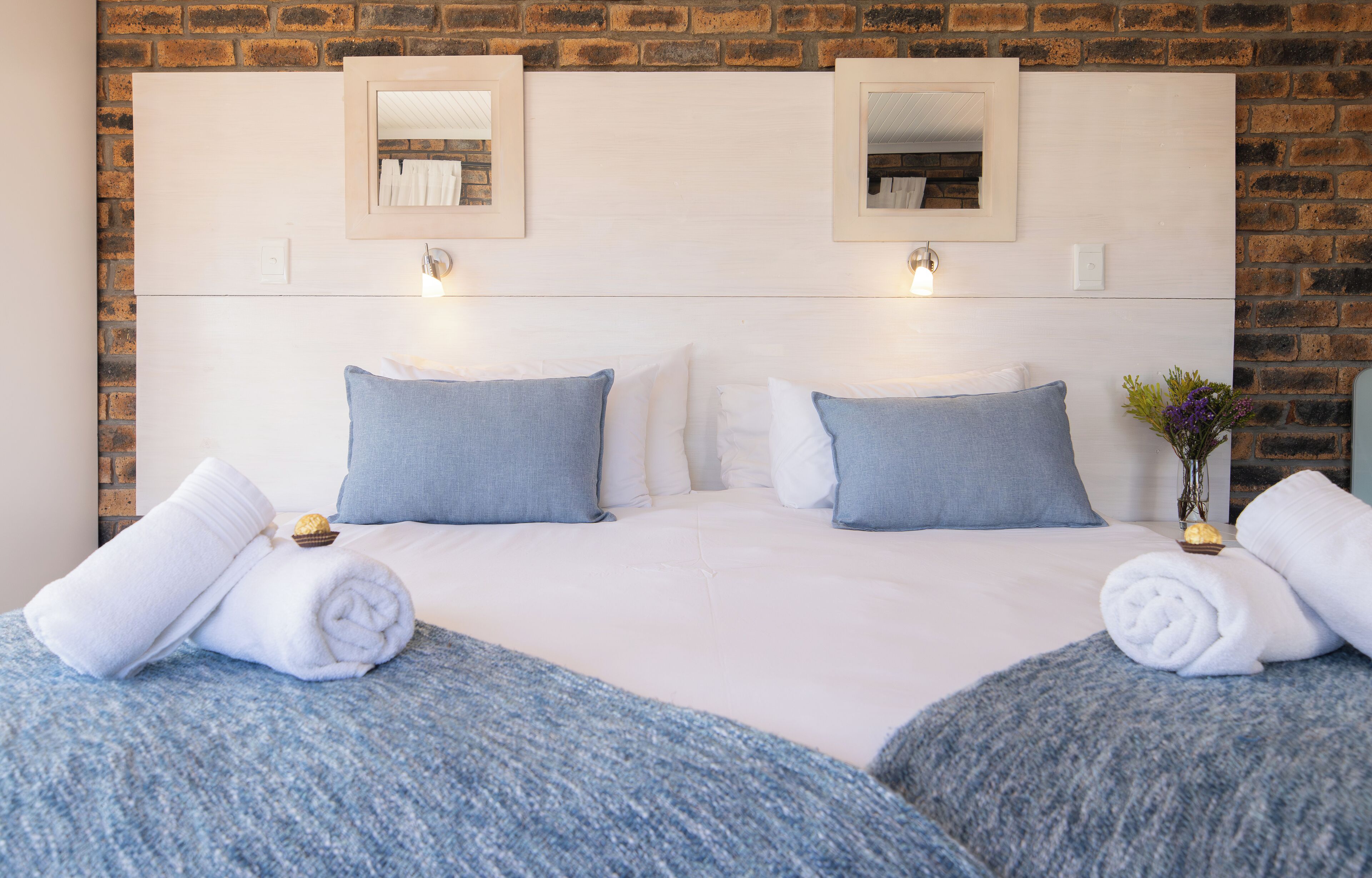 A double bed with white and blue bedding, two wall-mounted mirrors, and a small vase with flowers.