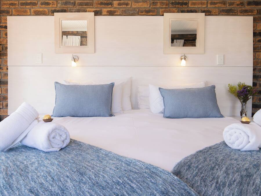 Una cama doble con ropa de cama blanca y azul, dos espejos en la pared y un jarrón con flores.