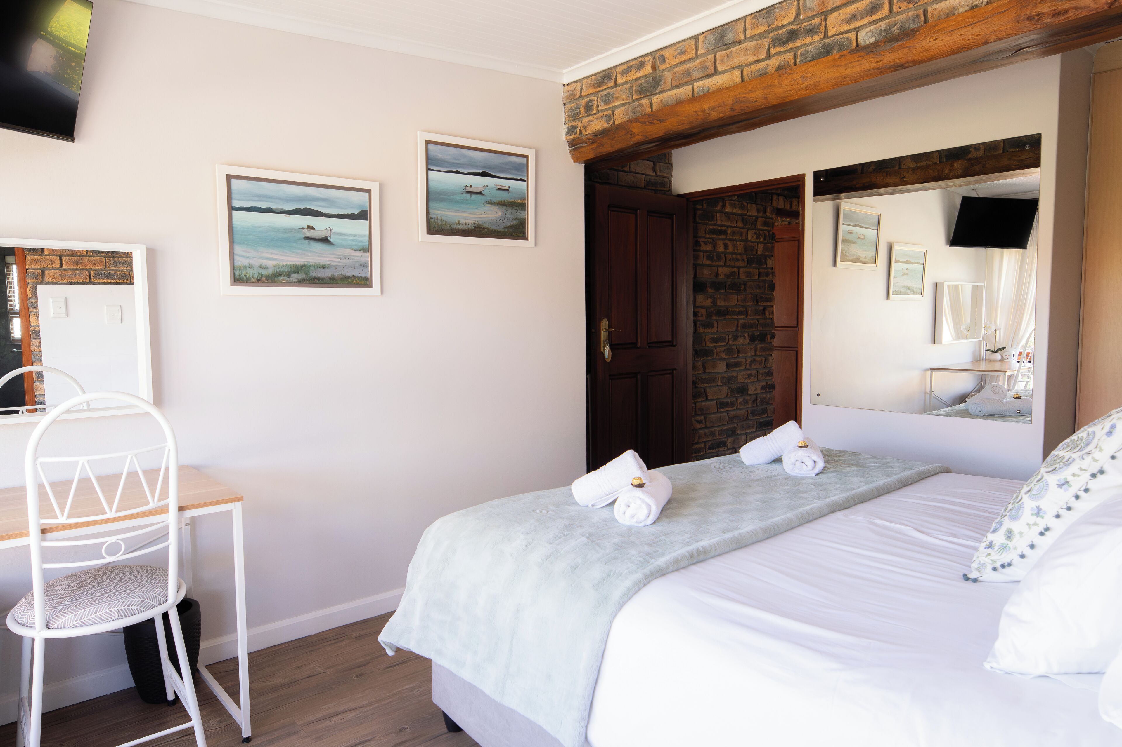 A bedroom with a bed, a desk, and a chair. There are two paintings on the wall and a mirror above the bed.