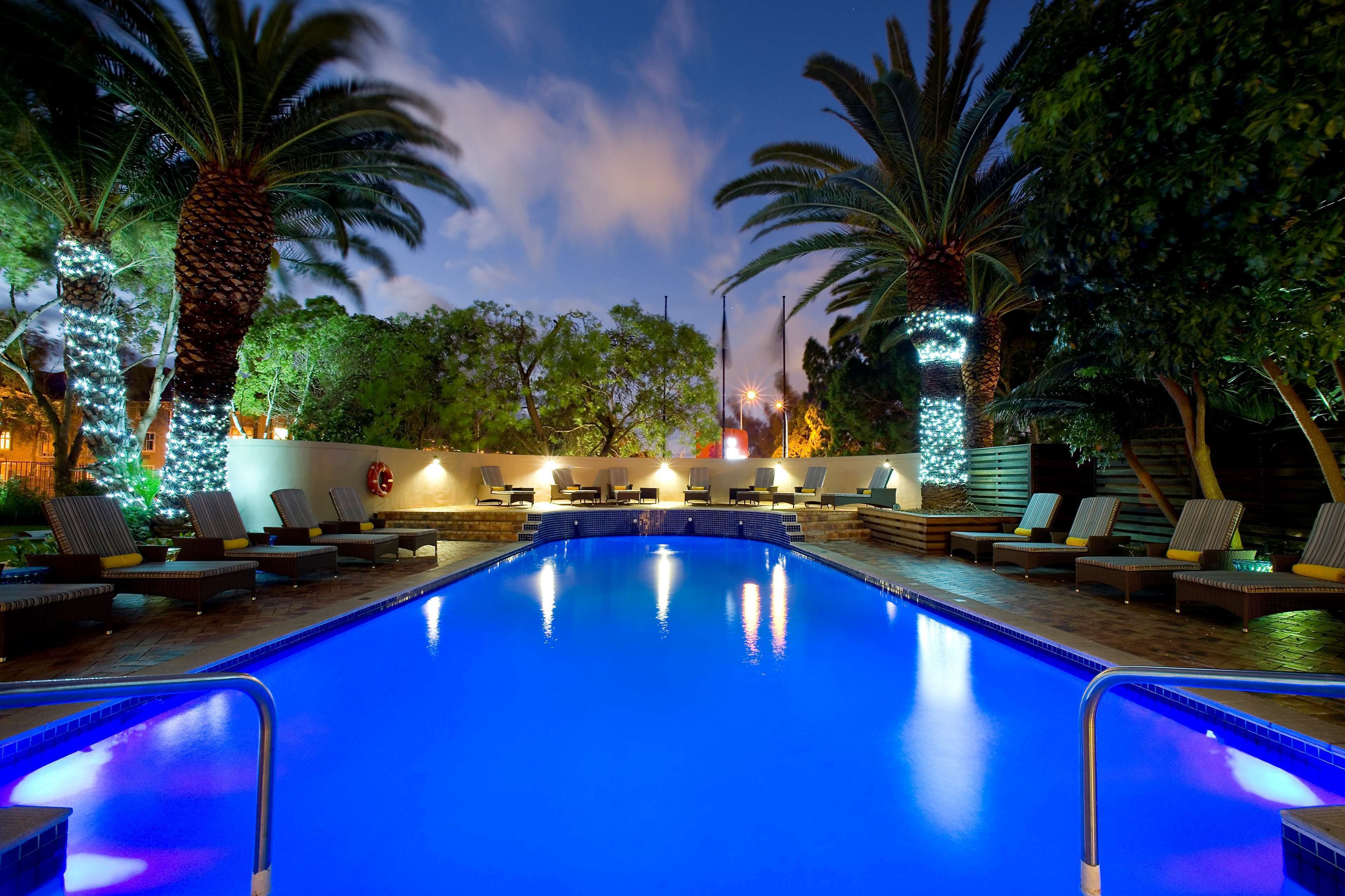 Une piscine entourée de chaises longues et de palmiers, le tout dans un espace extérieur bien éclairé.