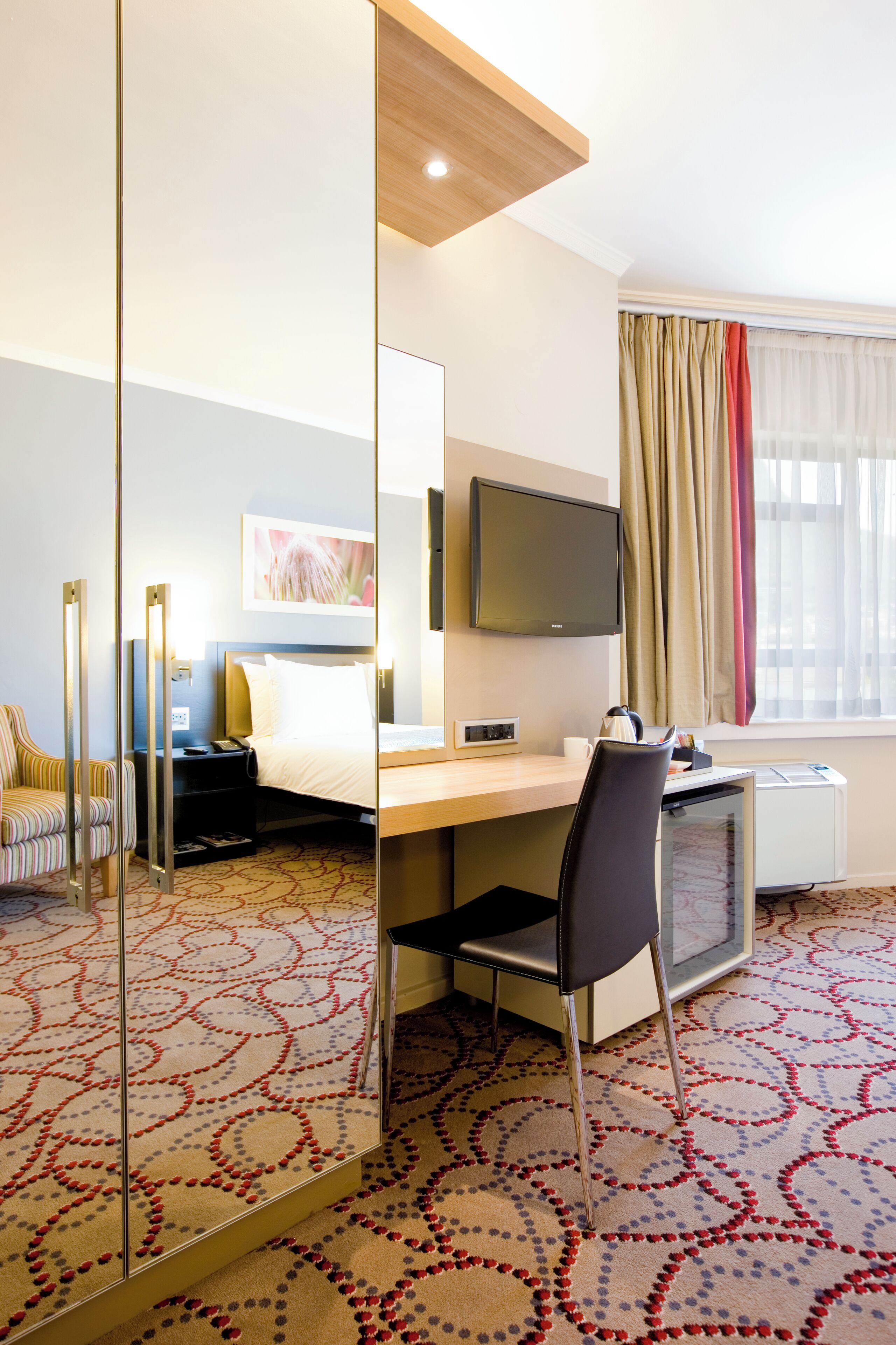 Habitación de hotel con cama, escritorio, silla, televisor y un espejo grande.