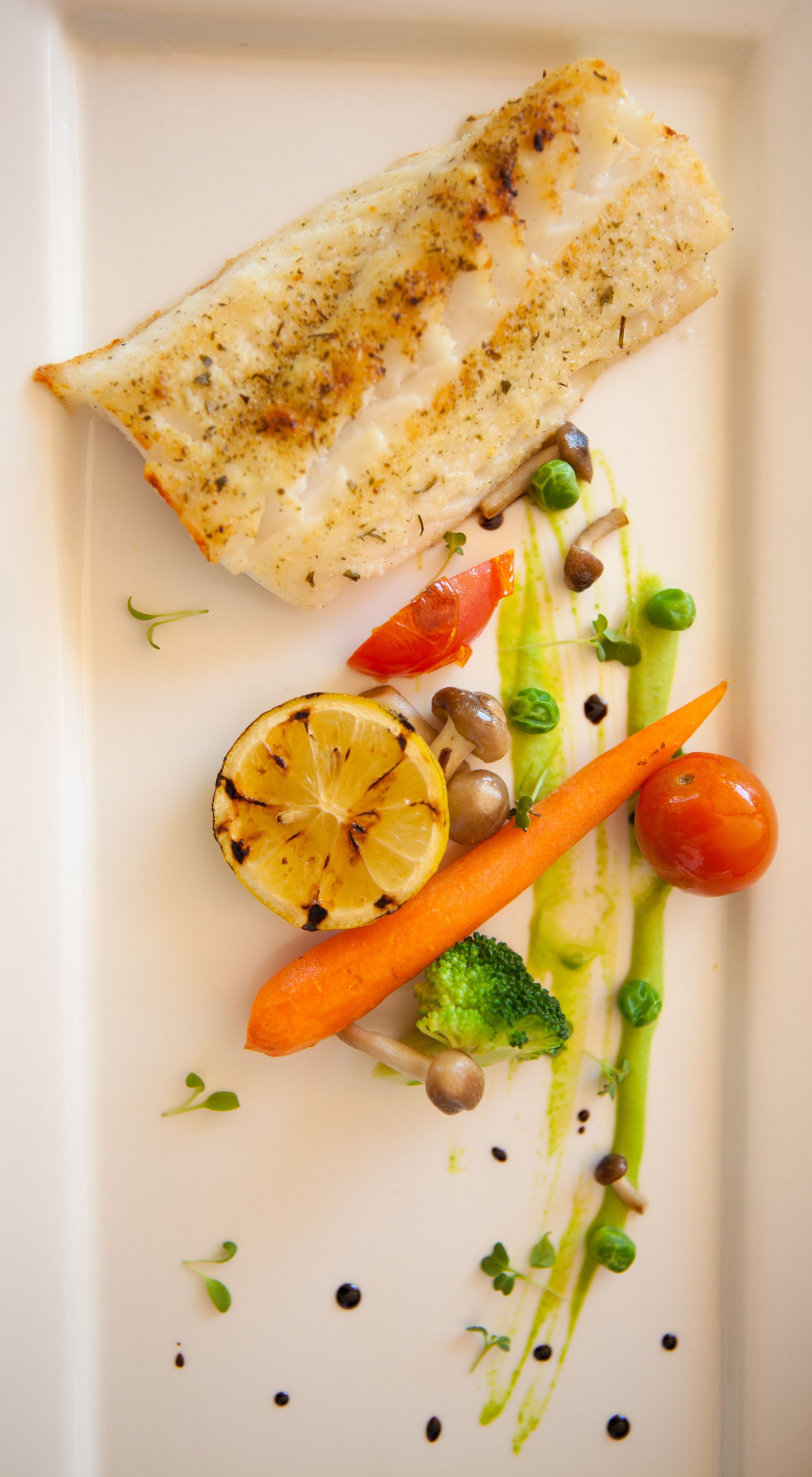 Une assiette blanche contenant un morceau de poisson grillé, un quartier de citron, des carottes, des brocolis et des tomates cerises.