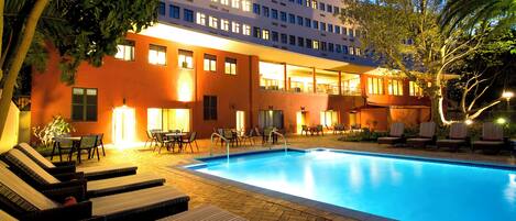 A hotel with a swimming pool, lounge chairs, and a well-lit building facade.