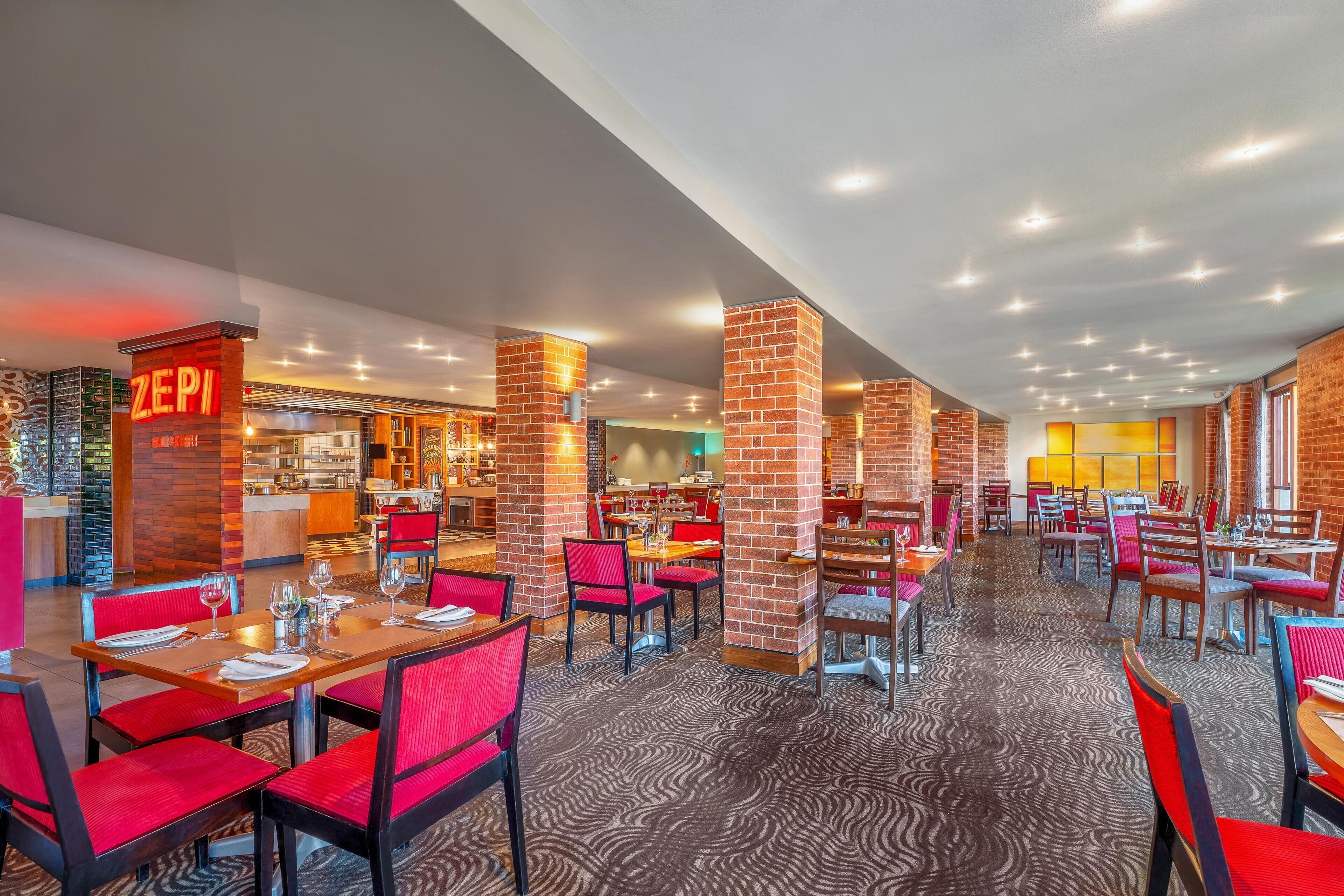 En rymlig hotellrestaurang med flera matplatser, röda stoppade stolar och tegelpelare.