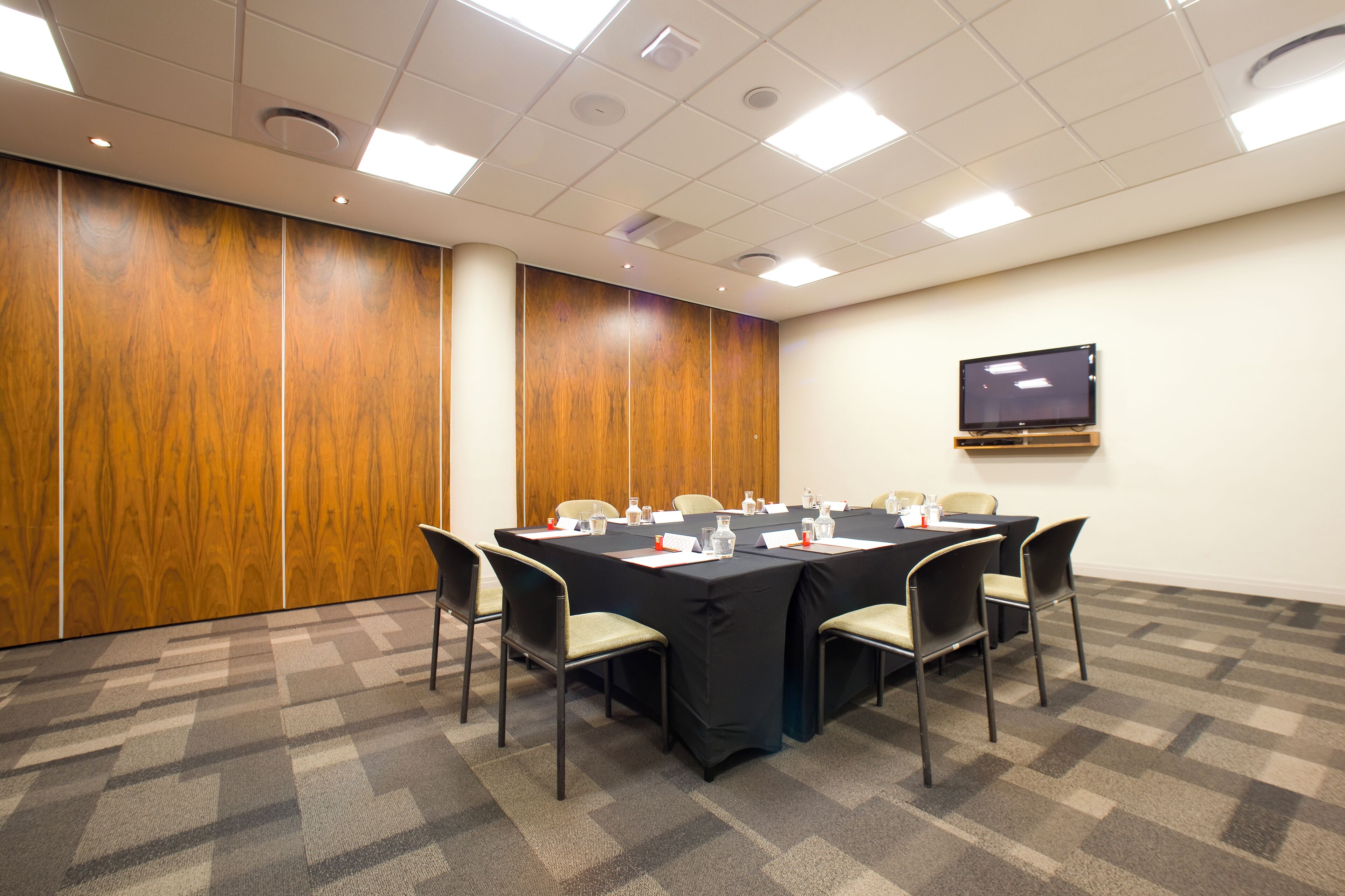 Un salón de conferencias con una larga mesa, sillas, un televisor de pantalla plana y un panel de pared de madera.