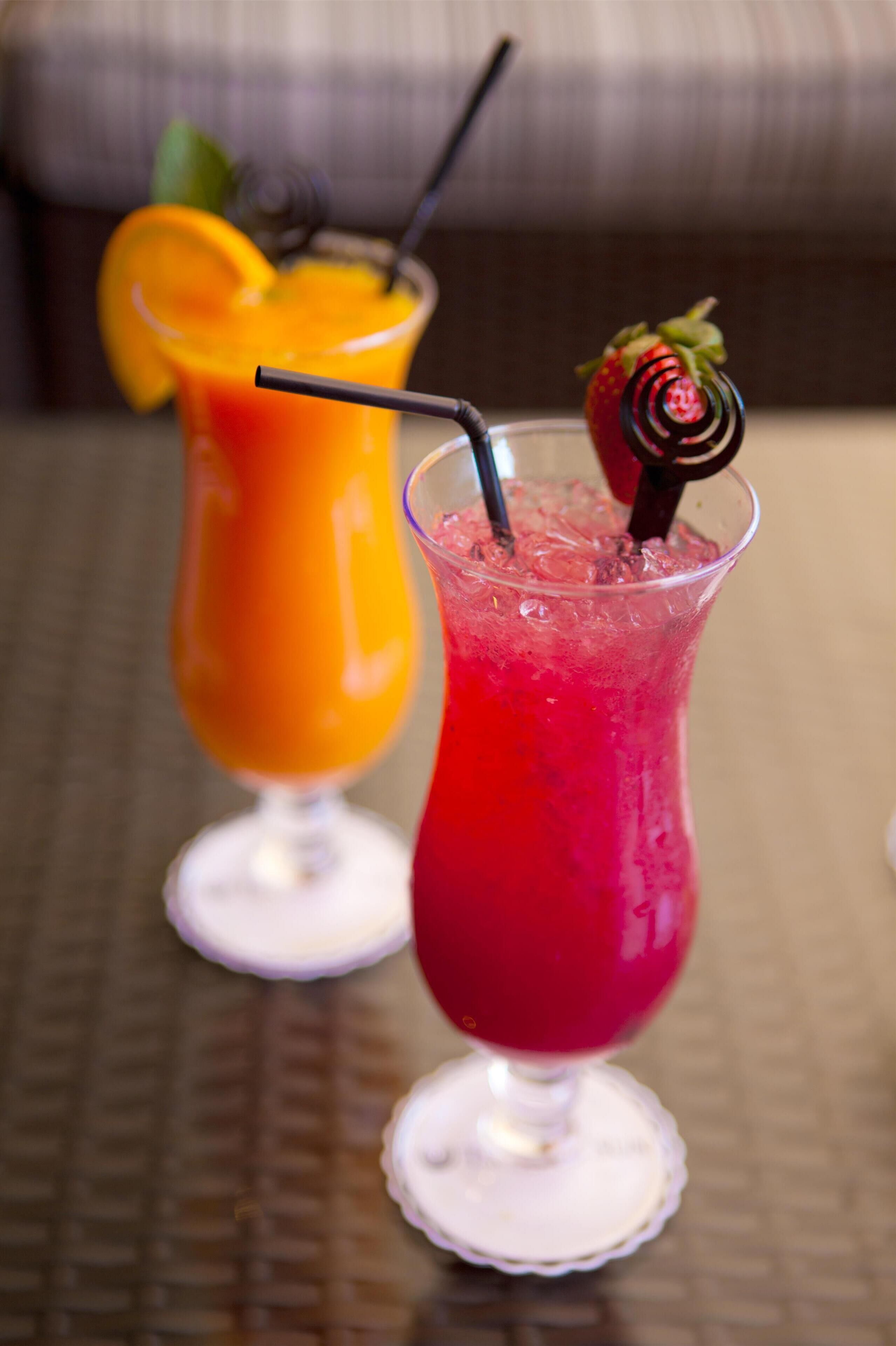 Two cocktails in glasses with straws and garnishes on a table.