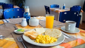 Une table dressée pour le petit-déjeuner, avec des toasts, des œufs brouillés, un verre de jus d’orange, une tasse de café et une bouteille d’eau.