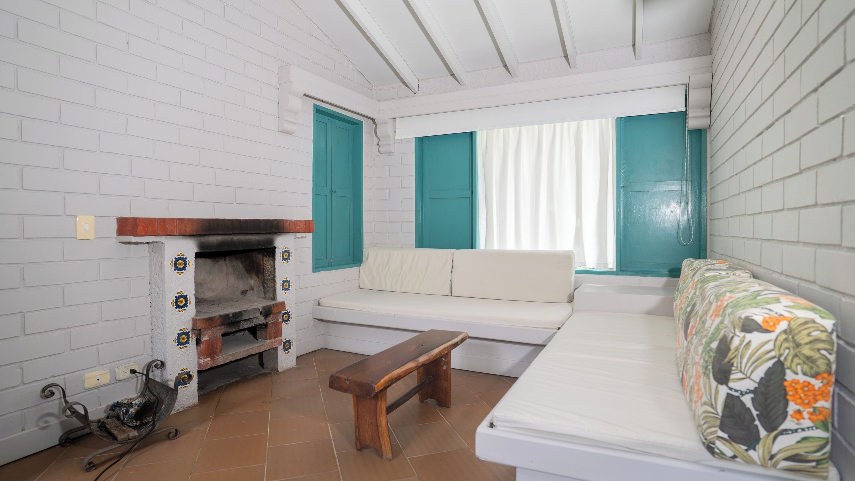 A small, cozy room with a white brick wall, a white sofa, a bench, and a fireplace with decorative tiles.