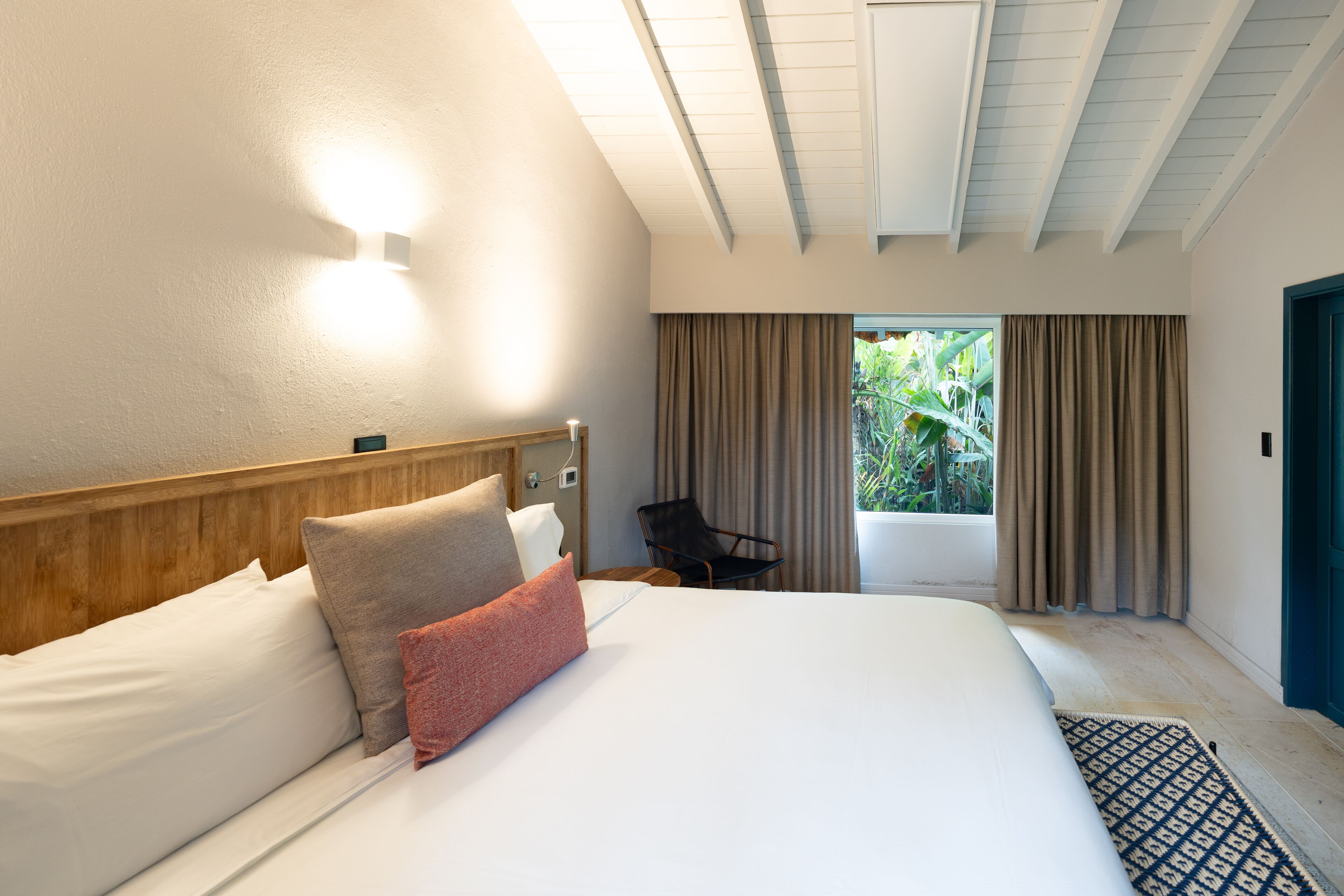 A hotel room with a large bed, a wooden headboard, a nightstand, a chair, and a view of greenery through the window.
