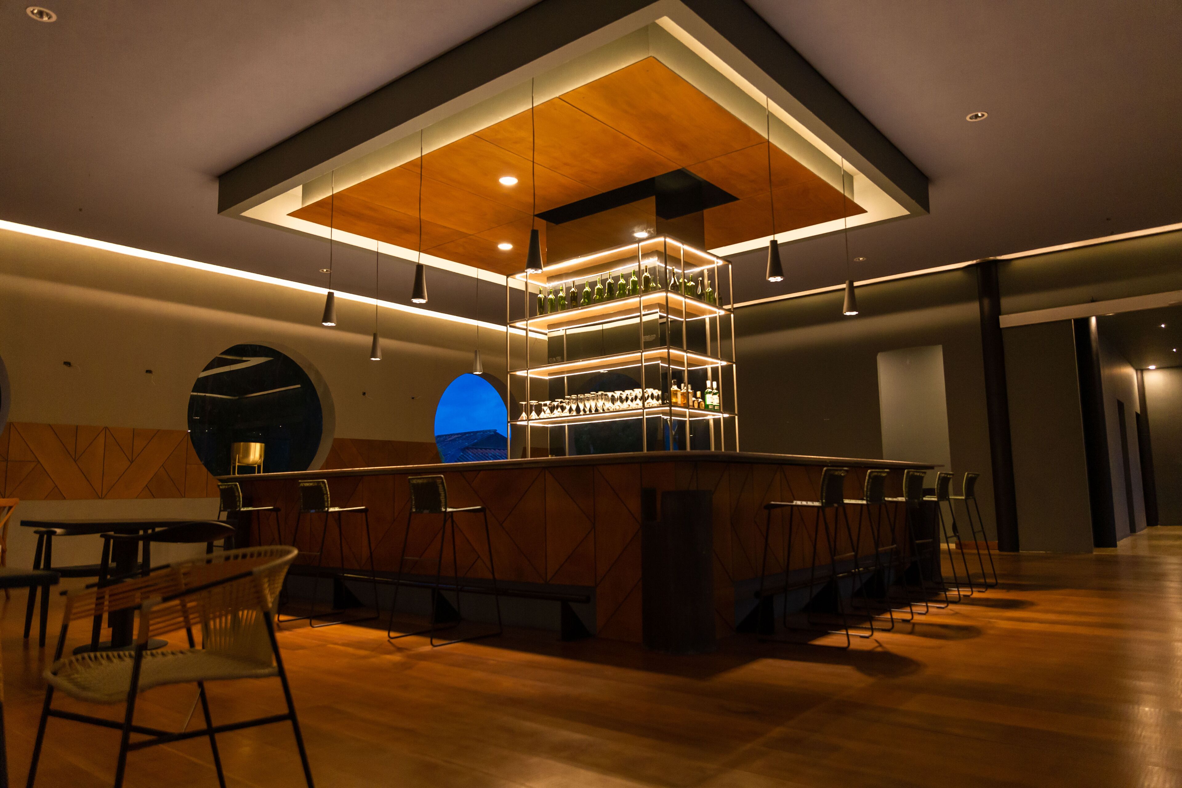 Un área de bar moderna con techo de madera, luces colgantes y una lámpara colgante azul. Hay taburetes de bar y una barra con botellas.