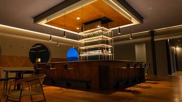 Un área de bar moderna con techo de madera, luces colgantes y una lámpara colgante azul. Hay taburetes de bar y una barra con botellas.