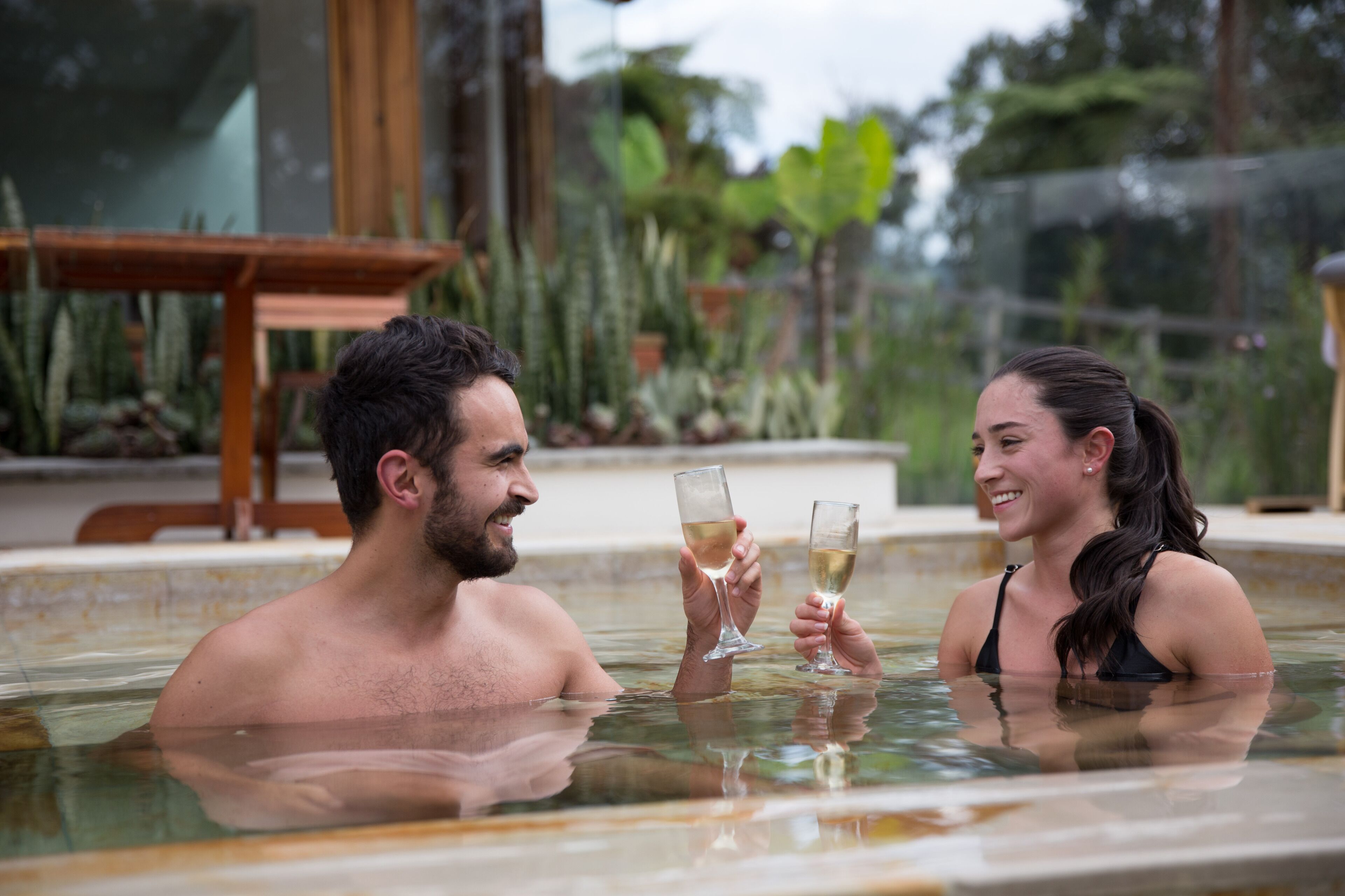 Dos personas en un jacuzzi con una mesa de madera y plantas al fondo.
