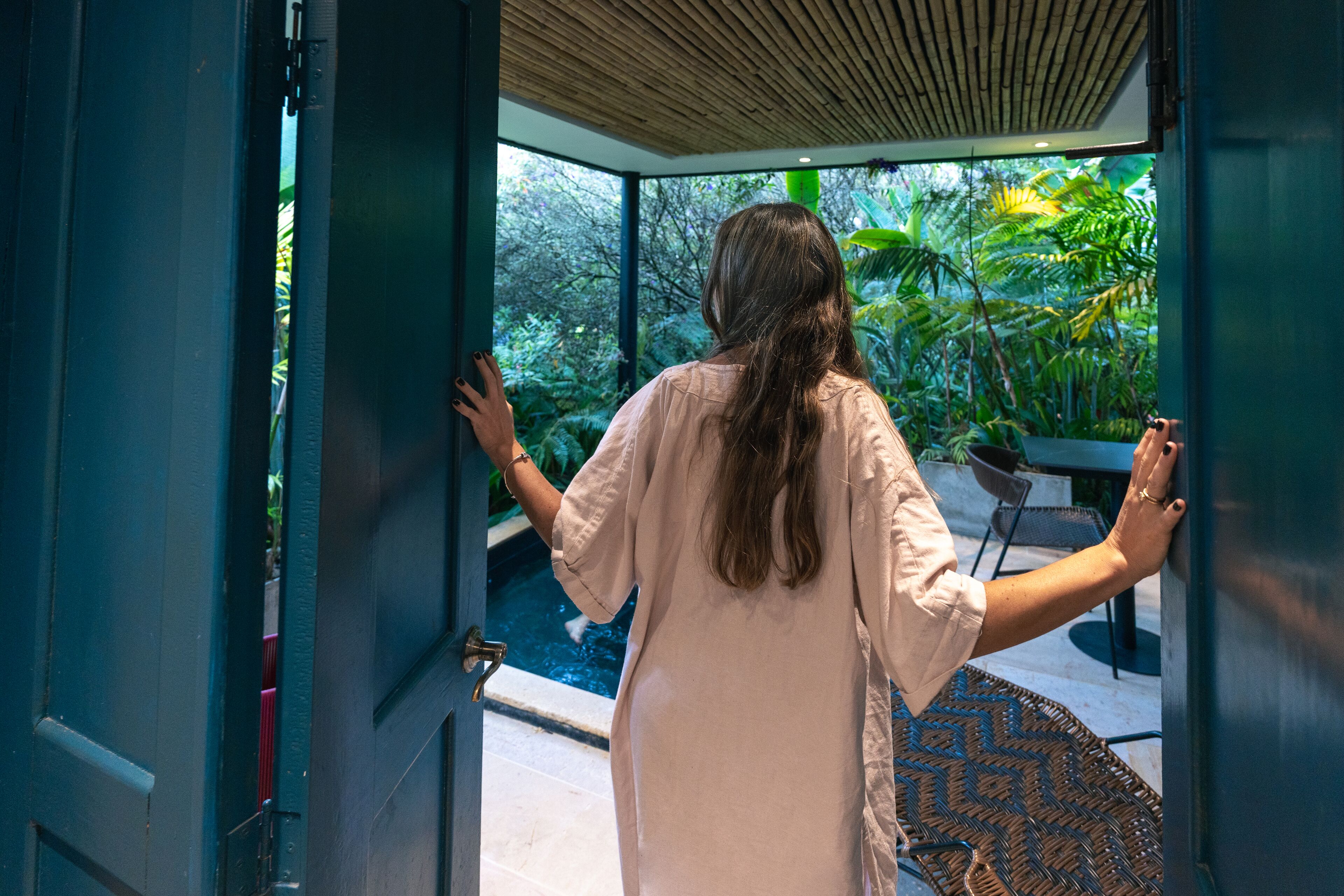 A woman standing at an open door, looking out at a lush garden with a pool and outdoor furniture.