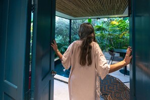 A woman standing at an open door, looking out at a lush garden with a pool and outdoor furniture. - Termales del Otoño (Villamaria)