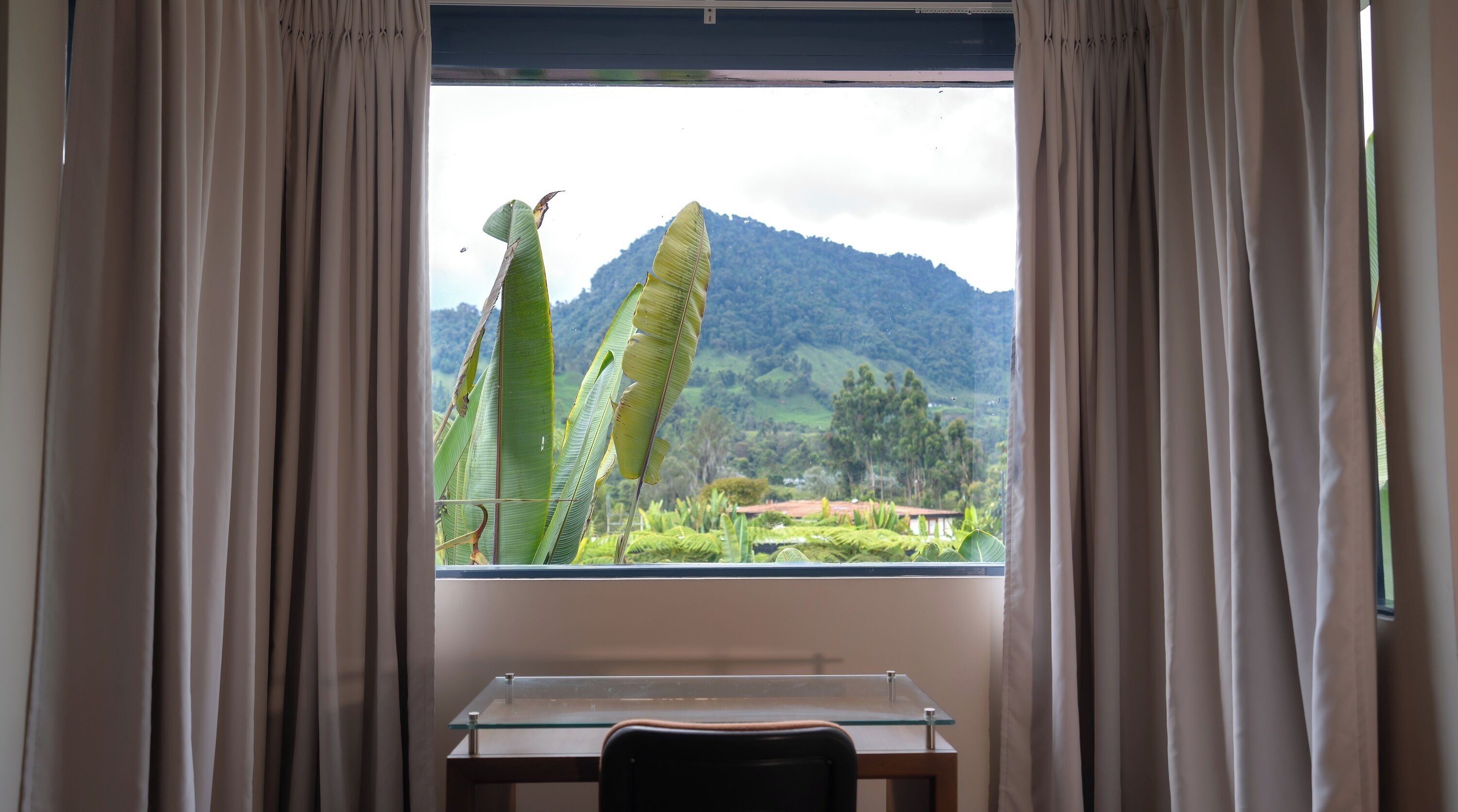 Quarto com escrivaninha e cadeira, janela com cortinas e vista para uma paisagem verde com montanhas.