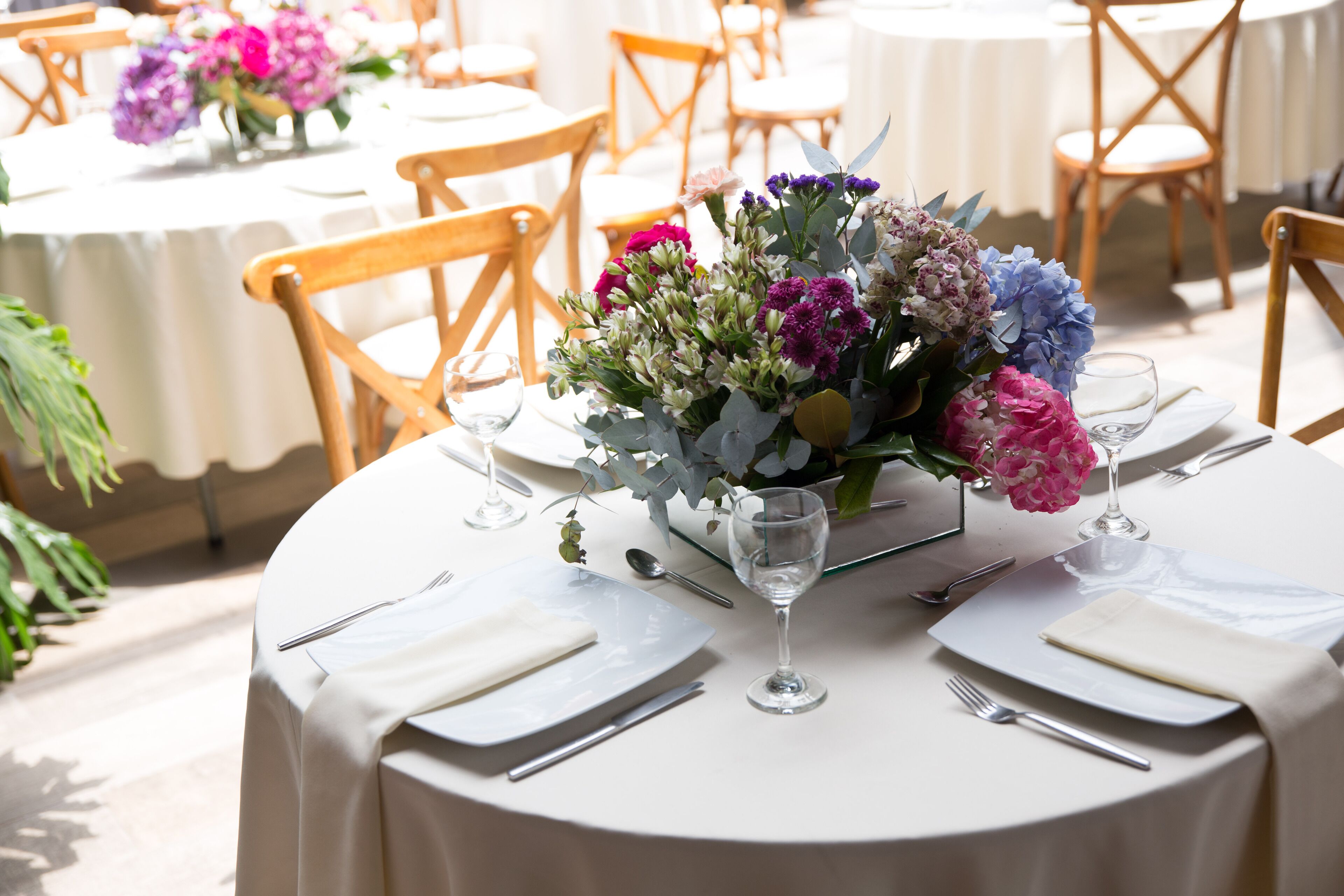 Una mesa redonda con mantel blanco, un florero de vidrio con flores y cubiertos colocados con esmero.