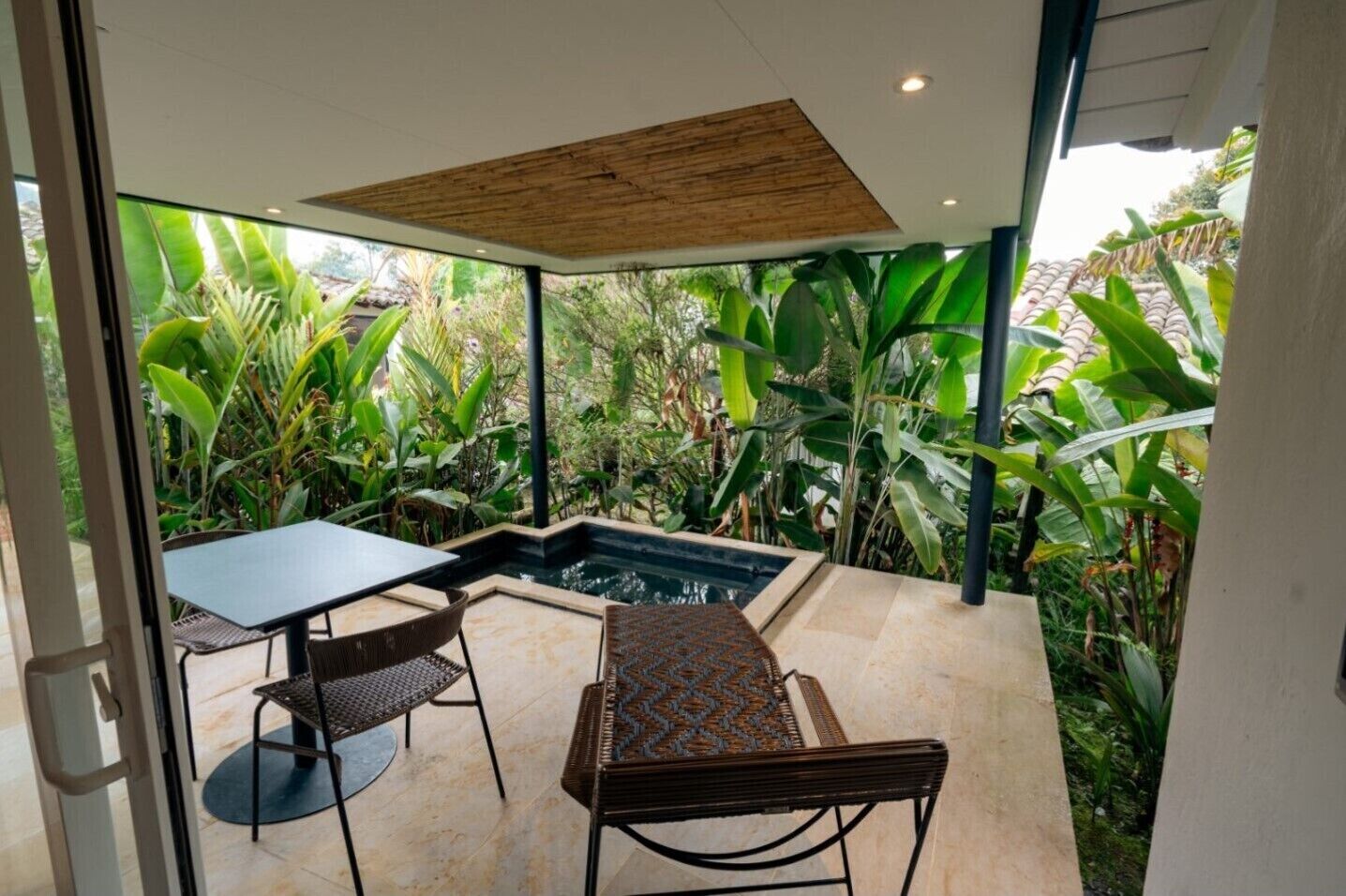 A modern outdoor patio with a pool, two tables, and chairs, surrounded by lush tropical plants.