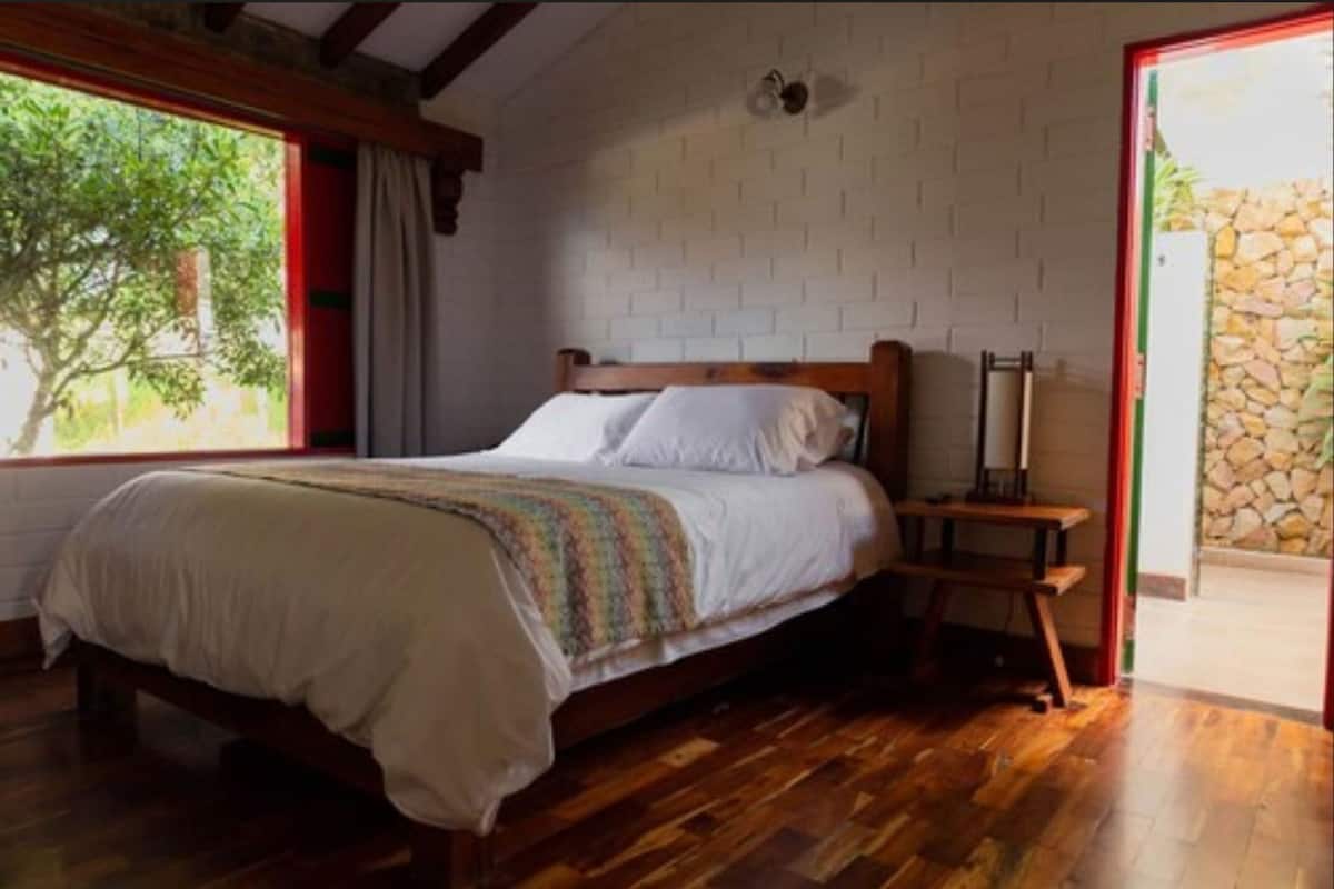 Ein Schlafzimmer mit einem hölzernen Bett, einem Nachttisch, einem Fenster mit Blick auf Grünflächen und einer Steinwand.
