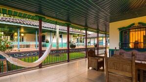 A covered patio area with a hammock, wooden furniture, and a view of a traditional building with tiled roofs and balconies. - Decameron Las Heliconias (Quimbaya)