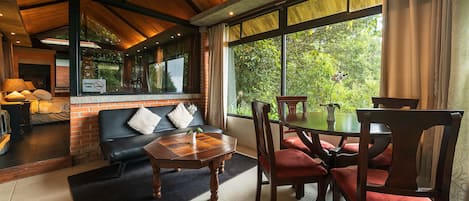 A cozy room with a fireplace, a sofa, a dining table with chairs, and large windows offering a view of greenery.