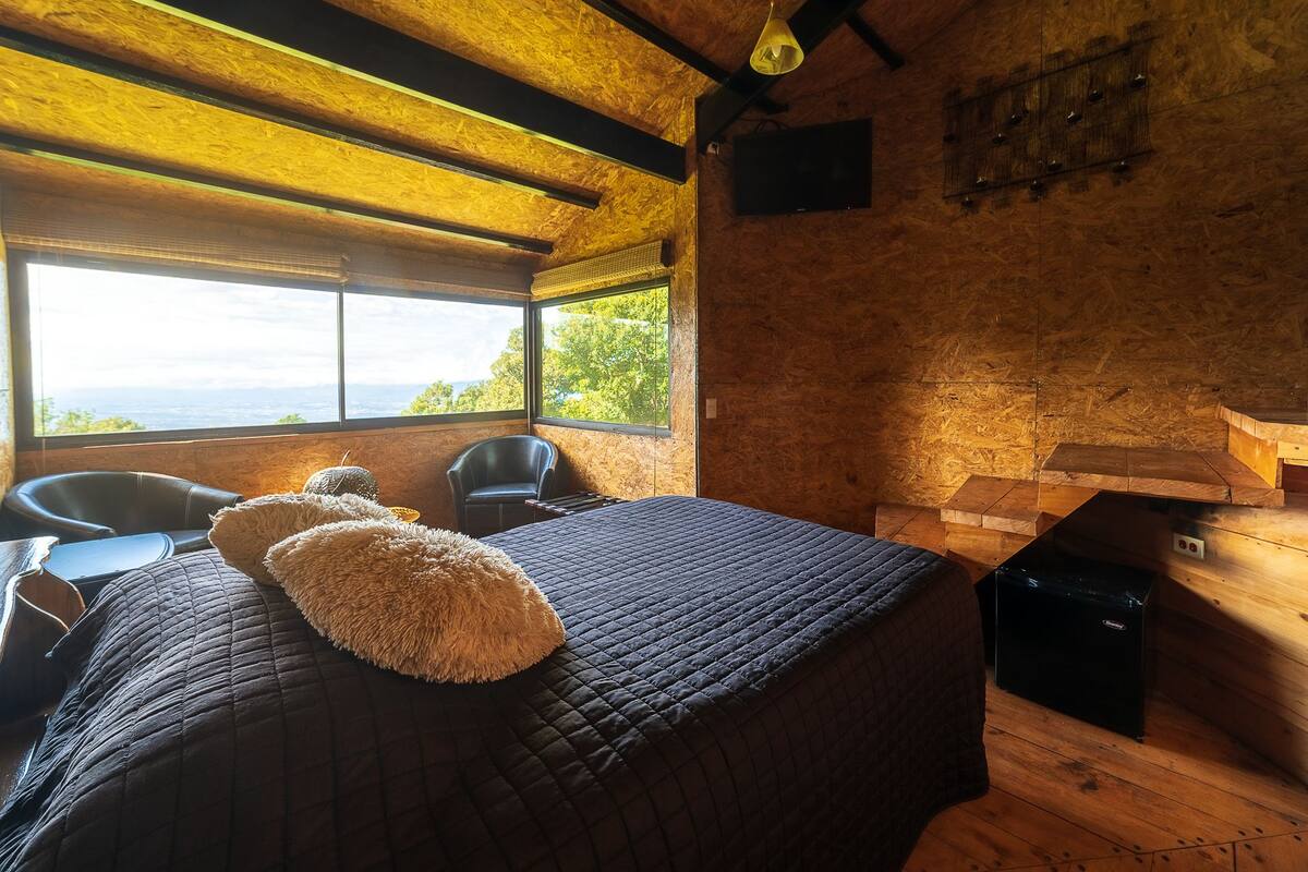 A cozy room with a large bed, two chairs, a wooden bench, and a view of the outdoors.