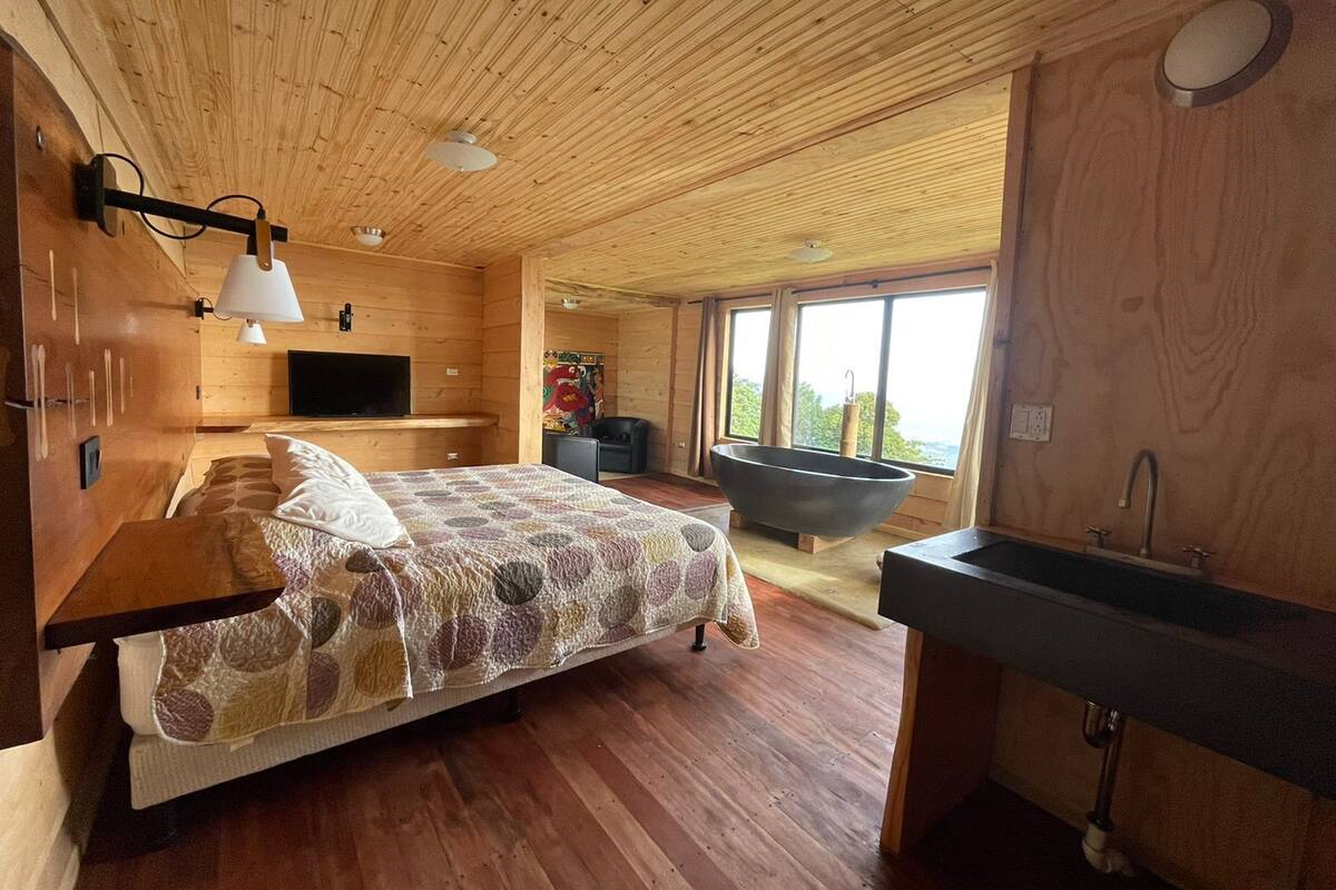 A wooden cabin bedroom with a bed, a large window, a bathtub, and a sink.