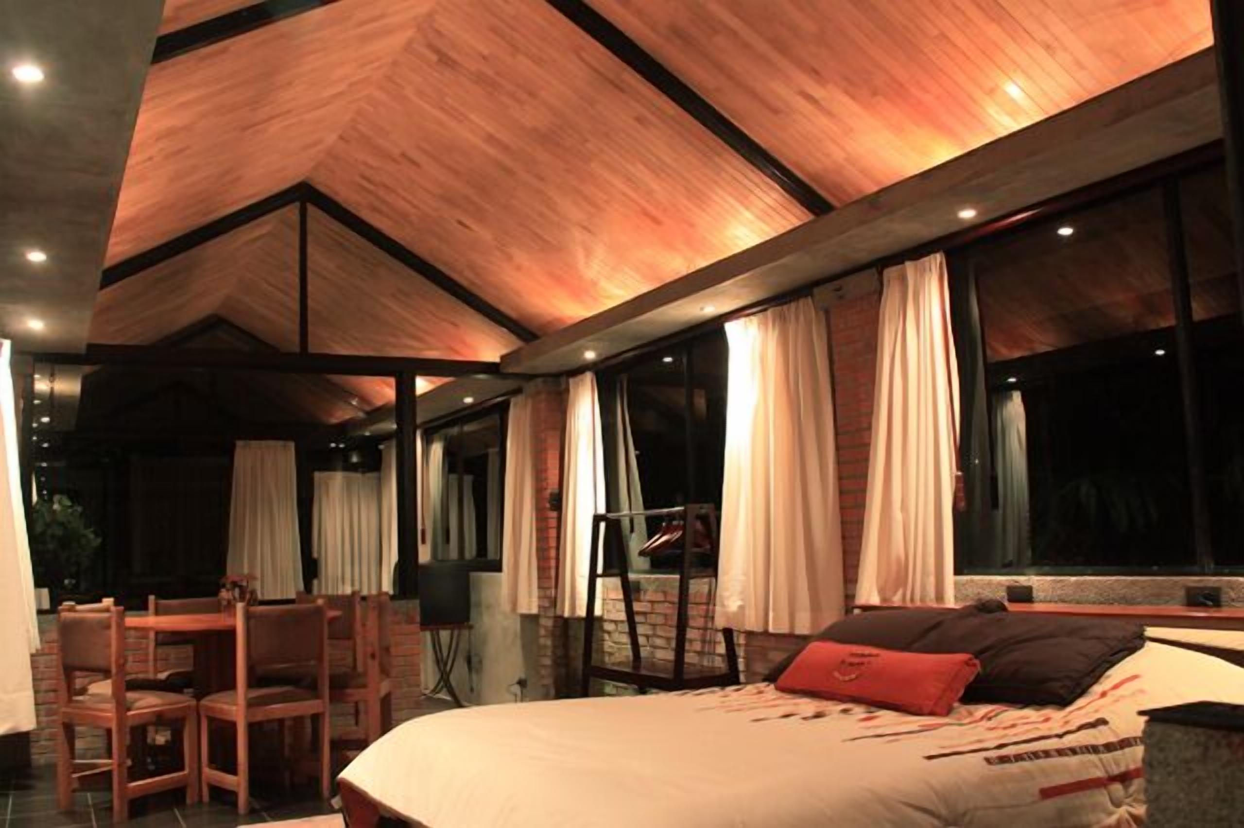 Habitación de hotel moderna con una cama grande, un escritorio y una silla. Amplias ventanas con cortinas y vistas al exterior.