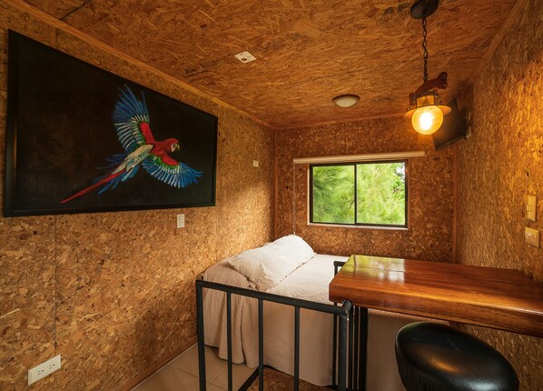 Una habitación con cama, escritorio, silla, ventana que ofrece vistas a follaje, y un cuadro de un loro.