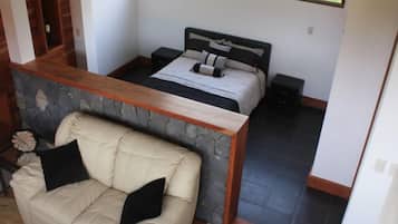 A bedroom with a stone accent wall, a bed with a headboard, a nightstand, a wooden bench, and a view of greenery outside.