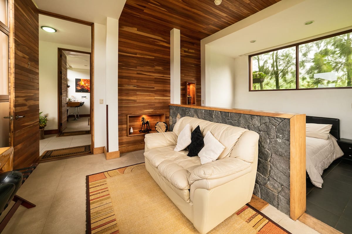 A modern living room with a beige sofa, a stone accent wall, and a large window offering a view of trees.