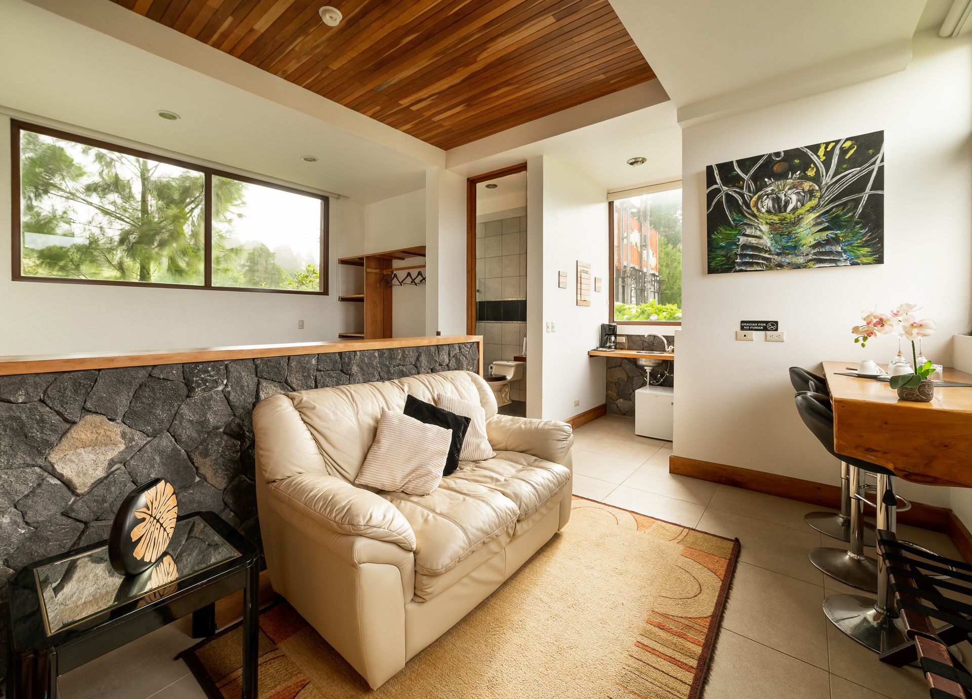 A modern living room with a stone accent wall, a beige sofa, a dining area with a wooden table, and a kitchen in the background.