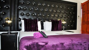 A bedroom with a tufted headboard, a bed with a purple blanket, two pillows, a remote control, and a nightstand with a lamp. - Lapologa Bed and Breakfast (Tzaneen)