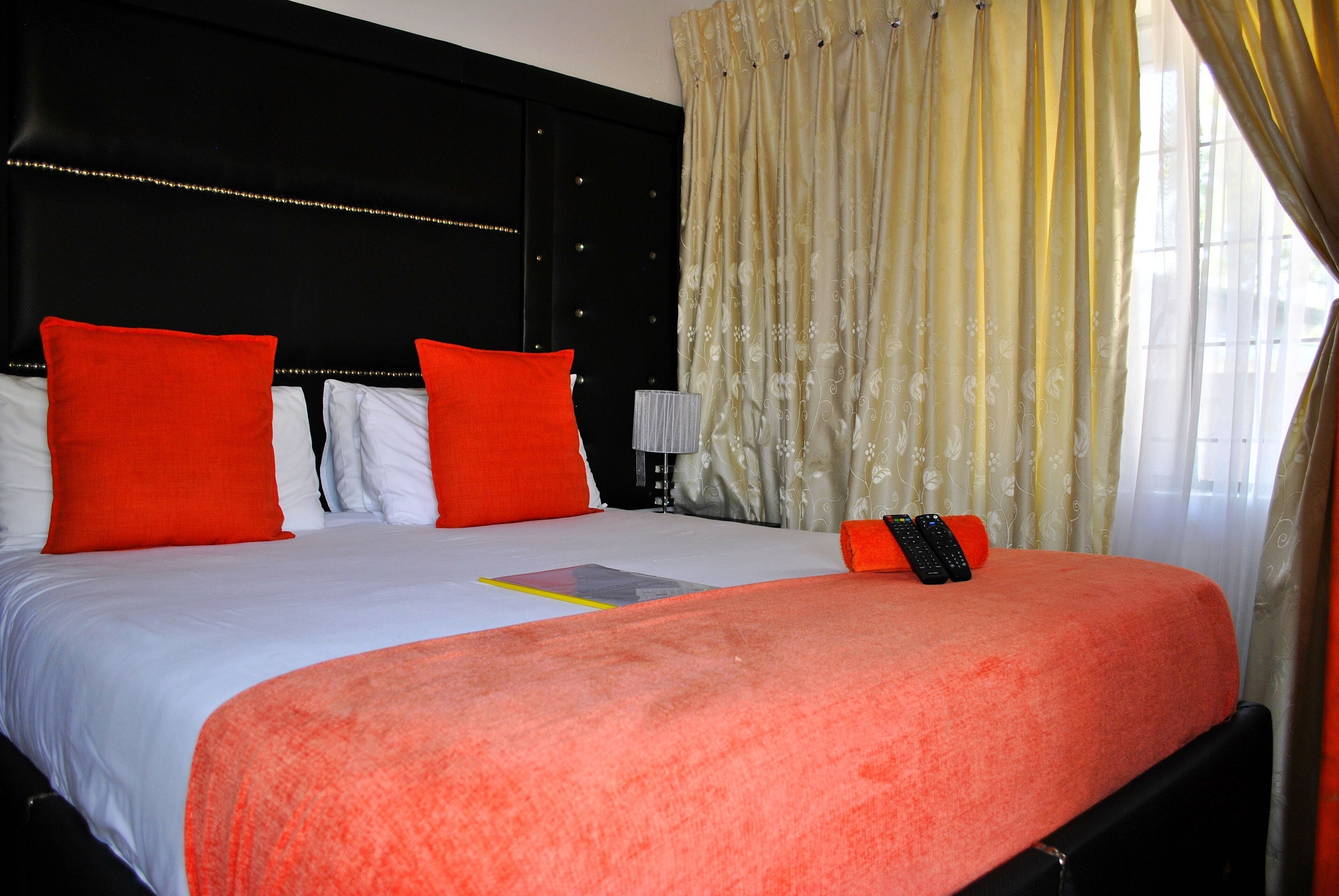 A hotel room with a black headboard, a bed with white and orange bedding, and a window with yellow curtains.