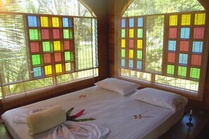 A room with a bed, a window with colorful stained glass, and a view of greenery outside.