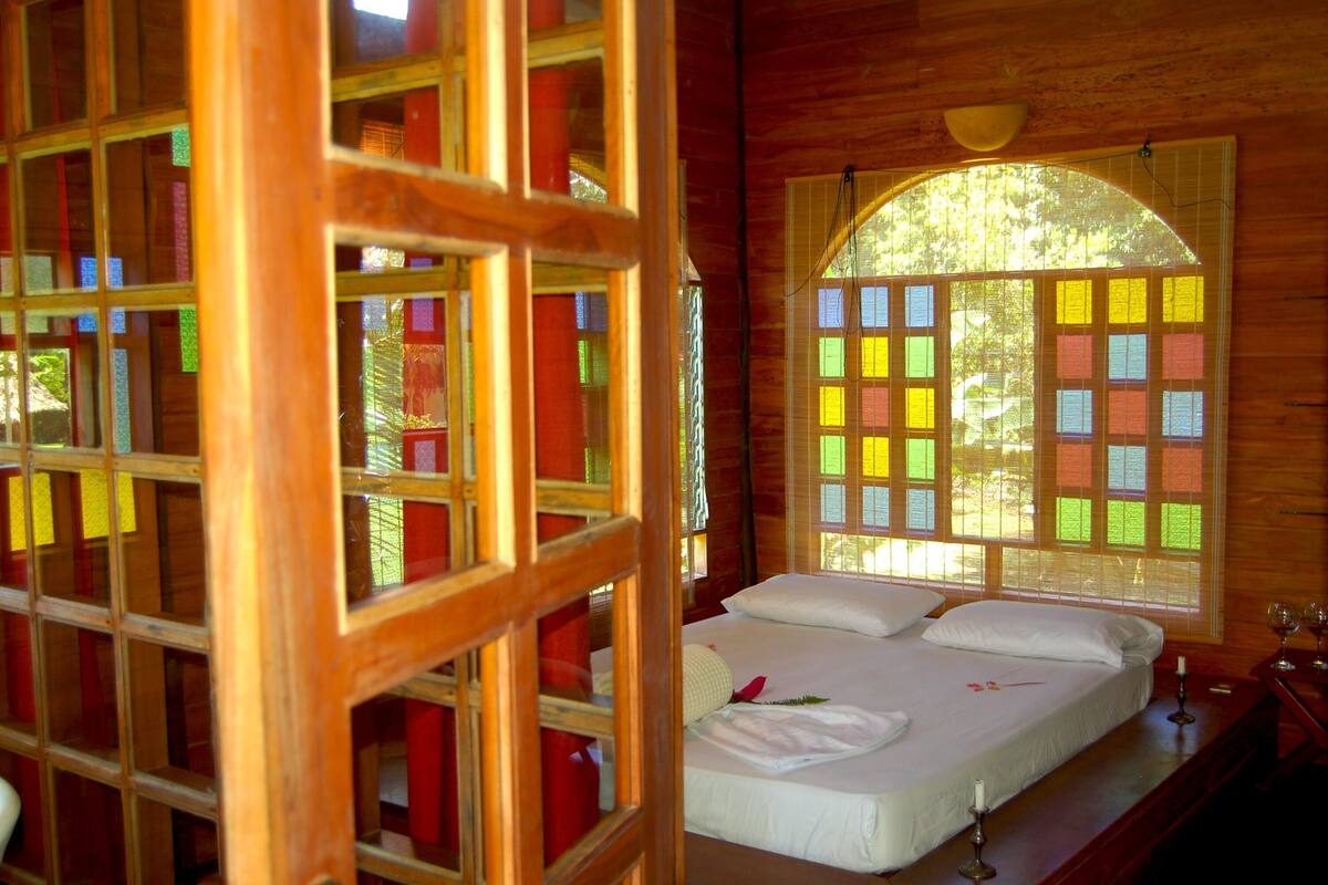 A wooden cabin room with a bed, a window with colorful notes, and a wooden partition.