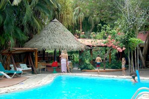 A pool area with lounge chairs, a thatched-roof structure, and lush greenery.