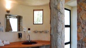 A bathroom with a wooden vanity, a stone pillar, a mirror, a window, and a wall-mounted soap dispenser. - Flying Crocodile (Sámara)