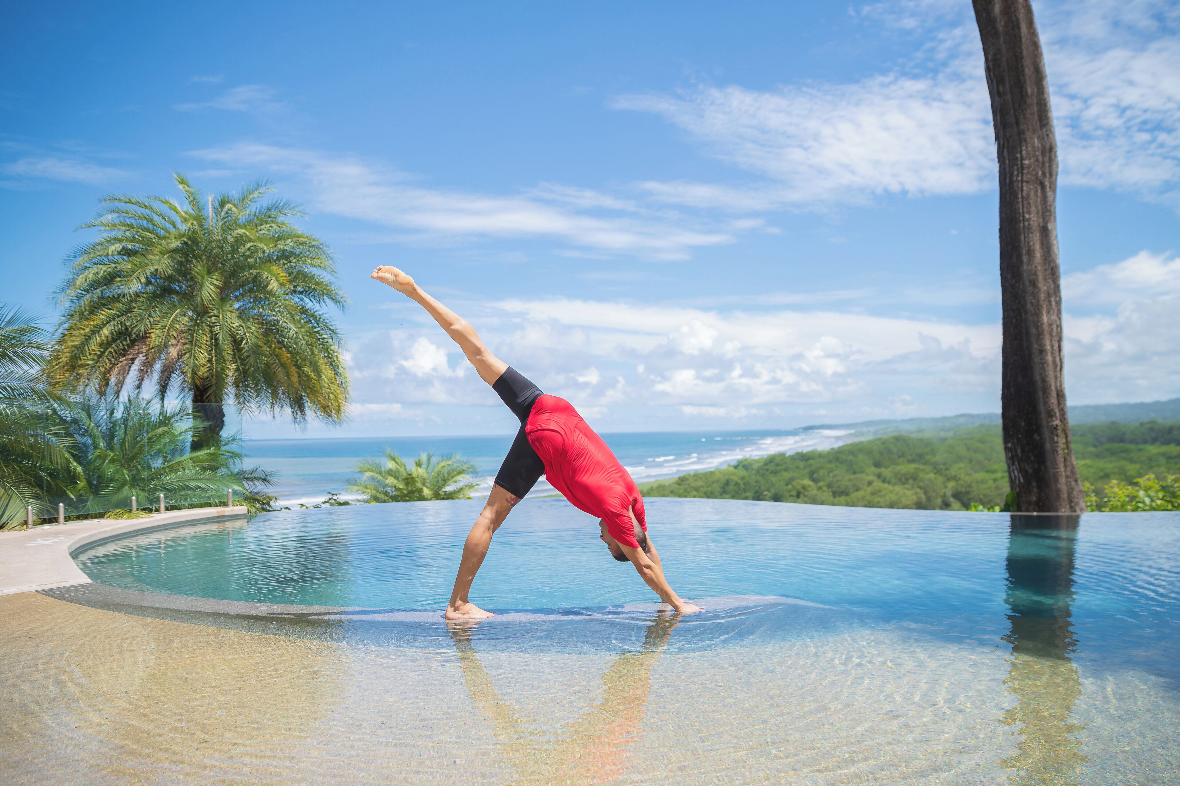 Una persona realiza una postura de yoga en el borde de una piscina cristalina con vista al océano y palmeras.