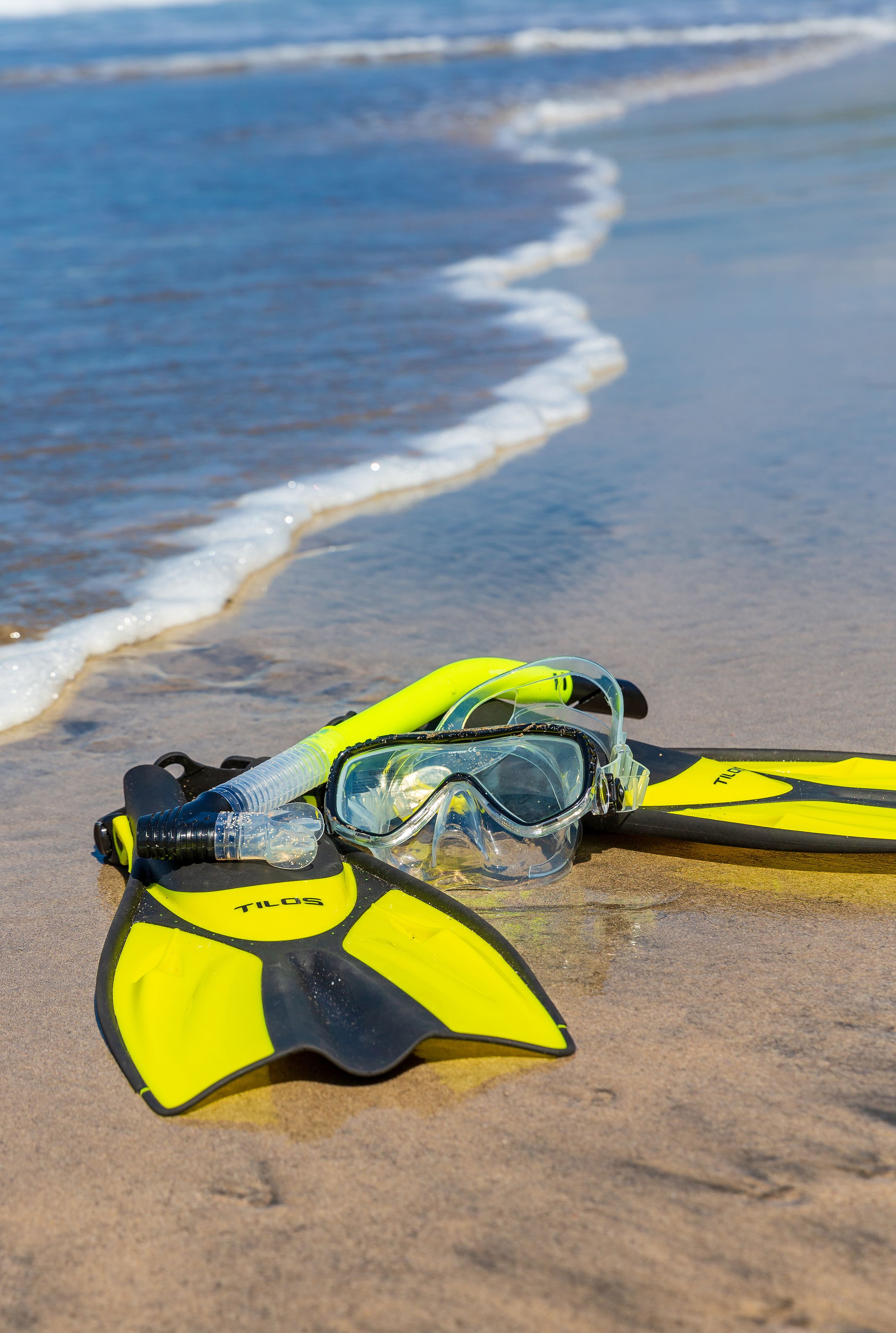 A pair of yellow and black flippers and a snorkel on a sandy beach with ocean waves in the background.