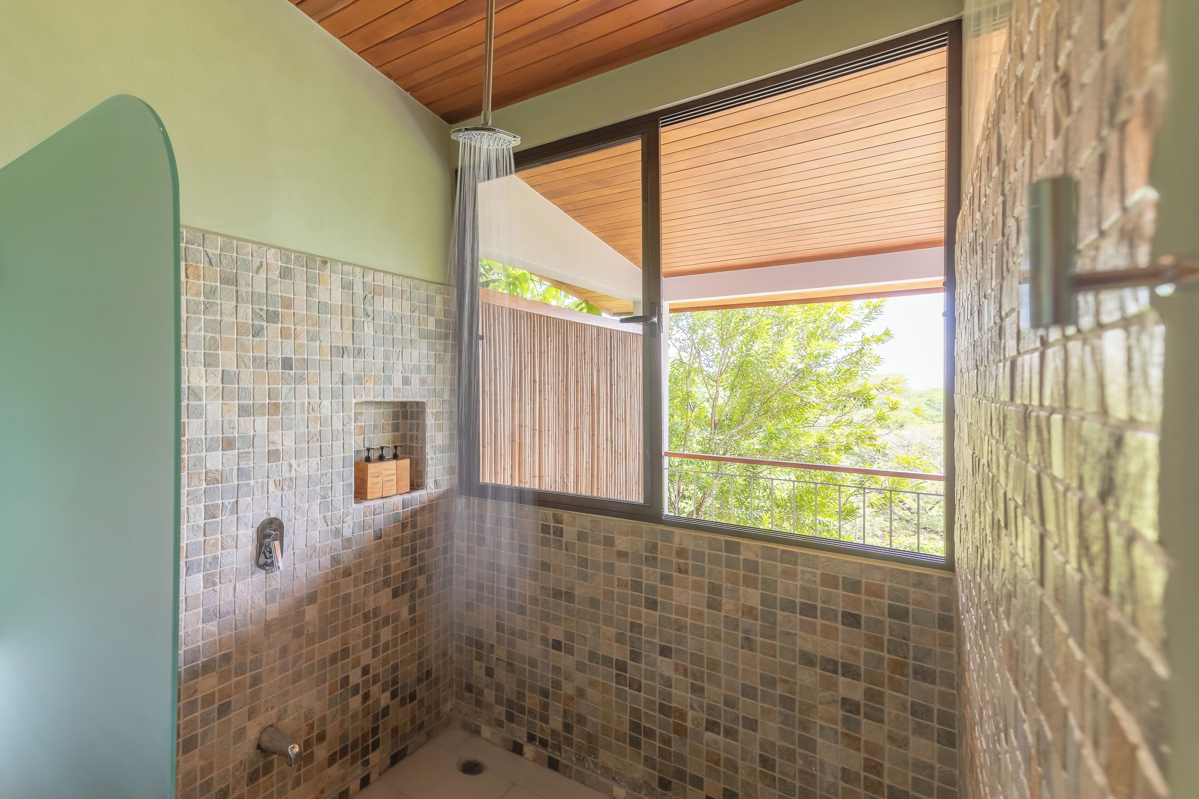 A bathroom with mosaic tile walls, a wooden ceiling, and a window with a view of greenery.