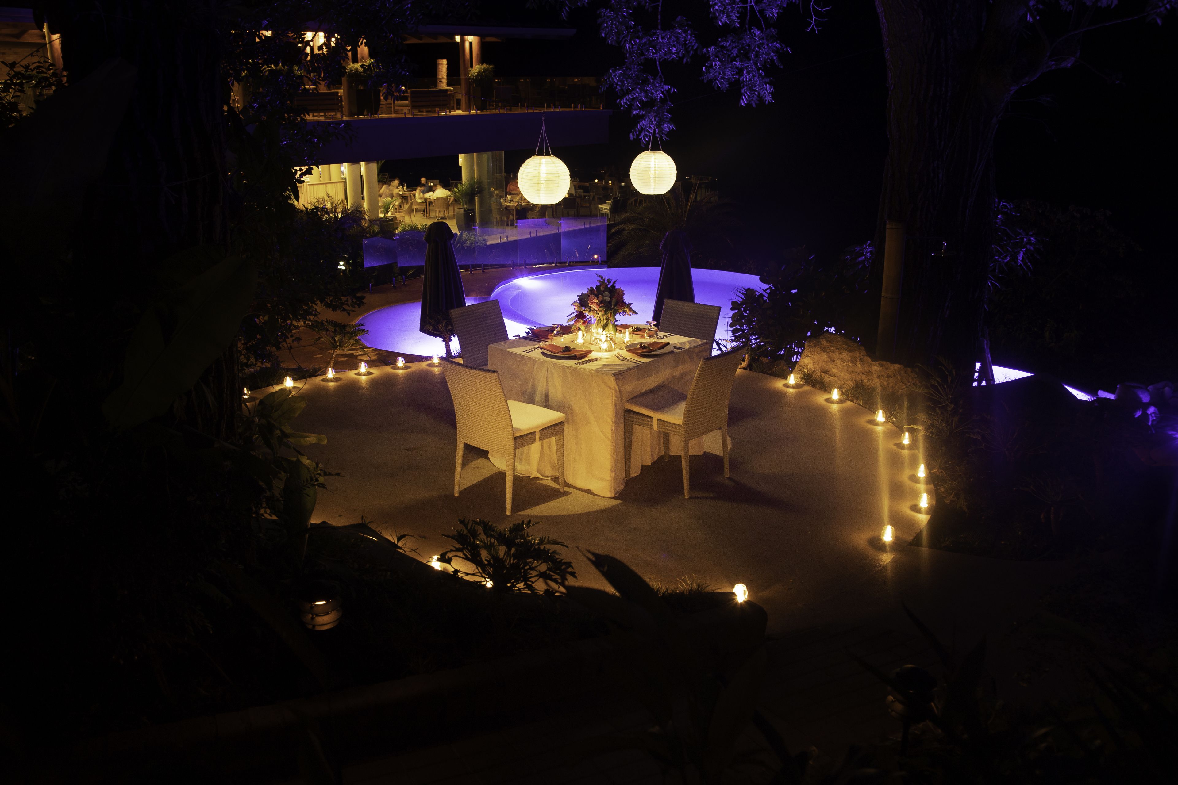 An outdoor dining area with a round table, chairs, and candles, surrounded by plants and trees.